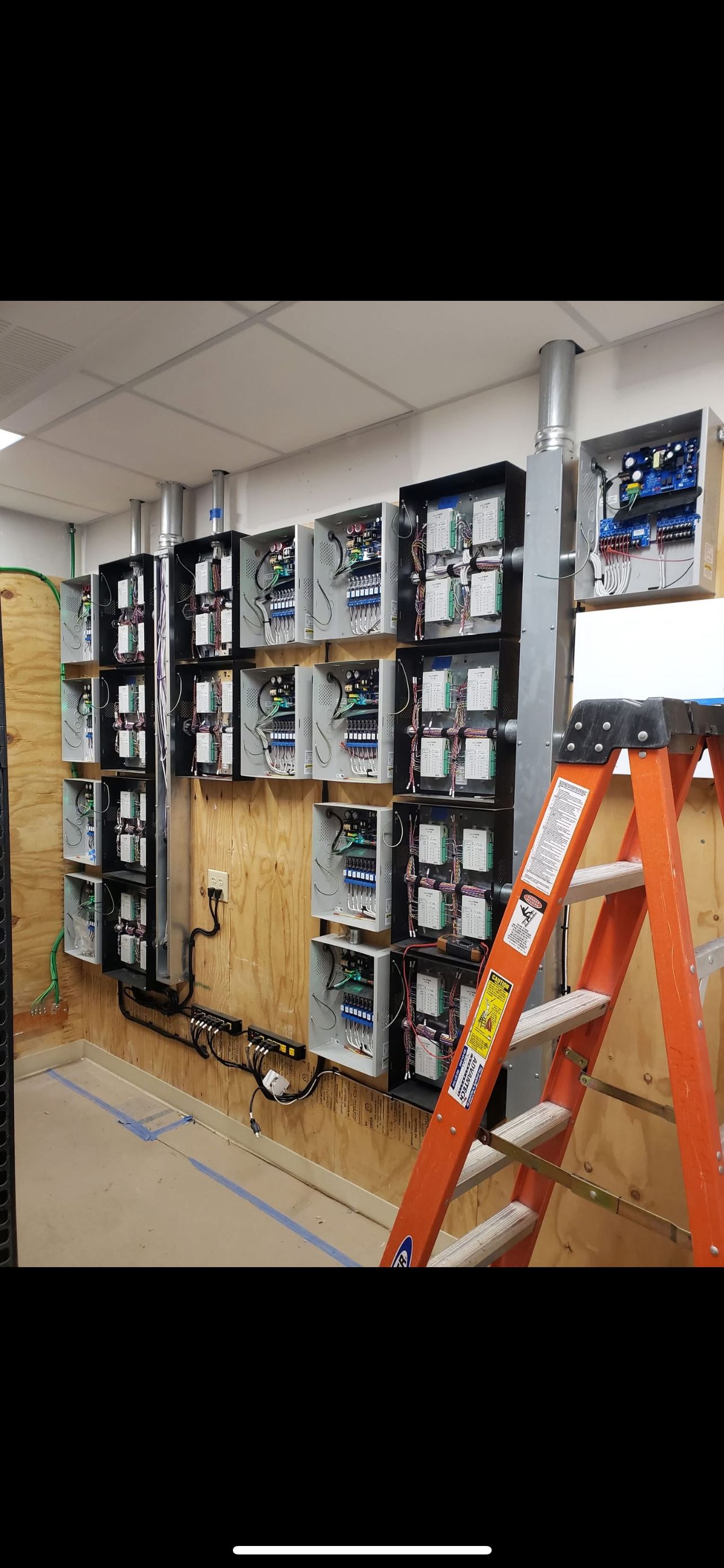 Electrical control panels with wiring and circuit boards on a wooden wall, next to an orange ladder.