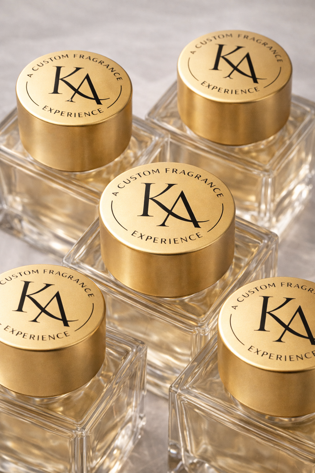 Glass perfume bottles with gold caps labeled "KA" and "A custom fragrance experience" stacked on a gray surface.