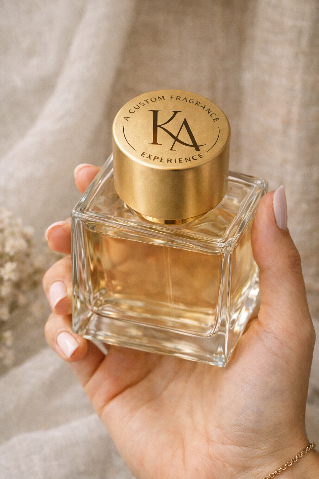 A hand holding a square glass perfume bottle with a gold cap that has the initials KA and the words "A Custom Fragrance Experience" printed on top.