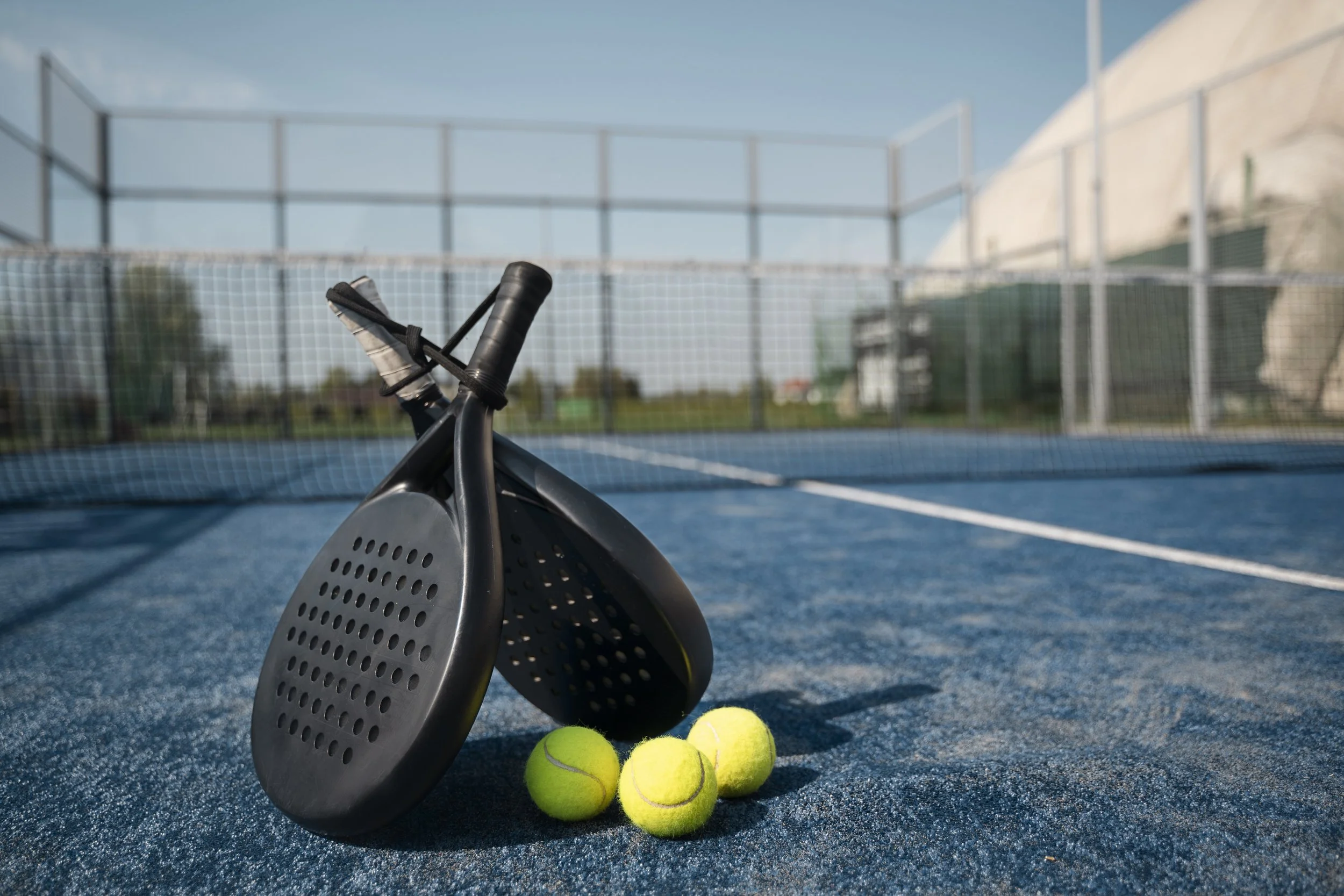 Tennis paddles and three tennis balls on a tennis court with a net and fence in the background.