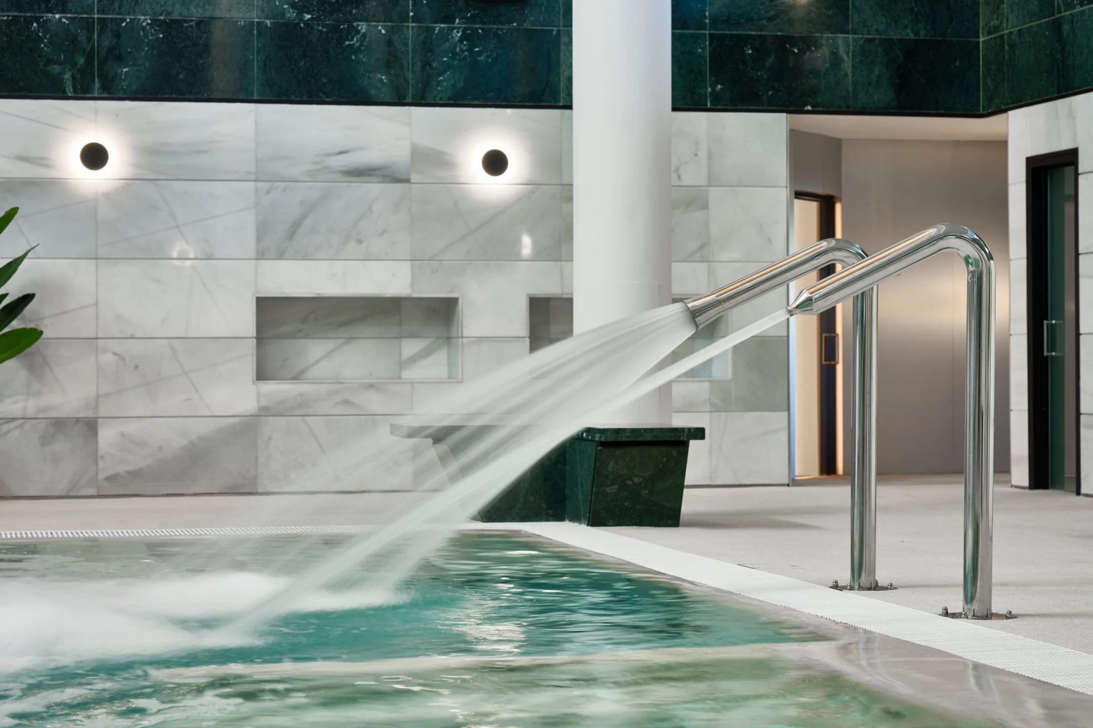 Indoor swimming pool with water flowing from two metallic spouts into the pool, surrounded by modern tiled walls and a white floor.