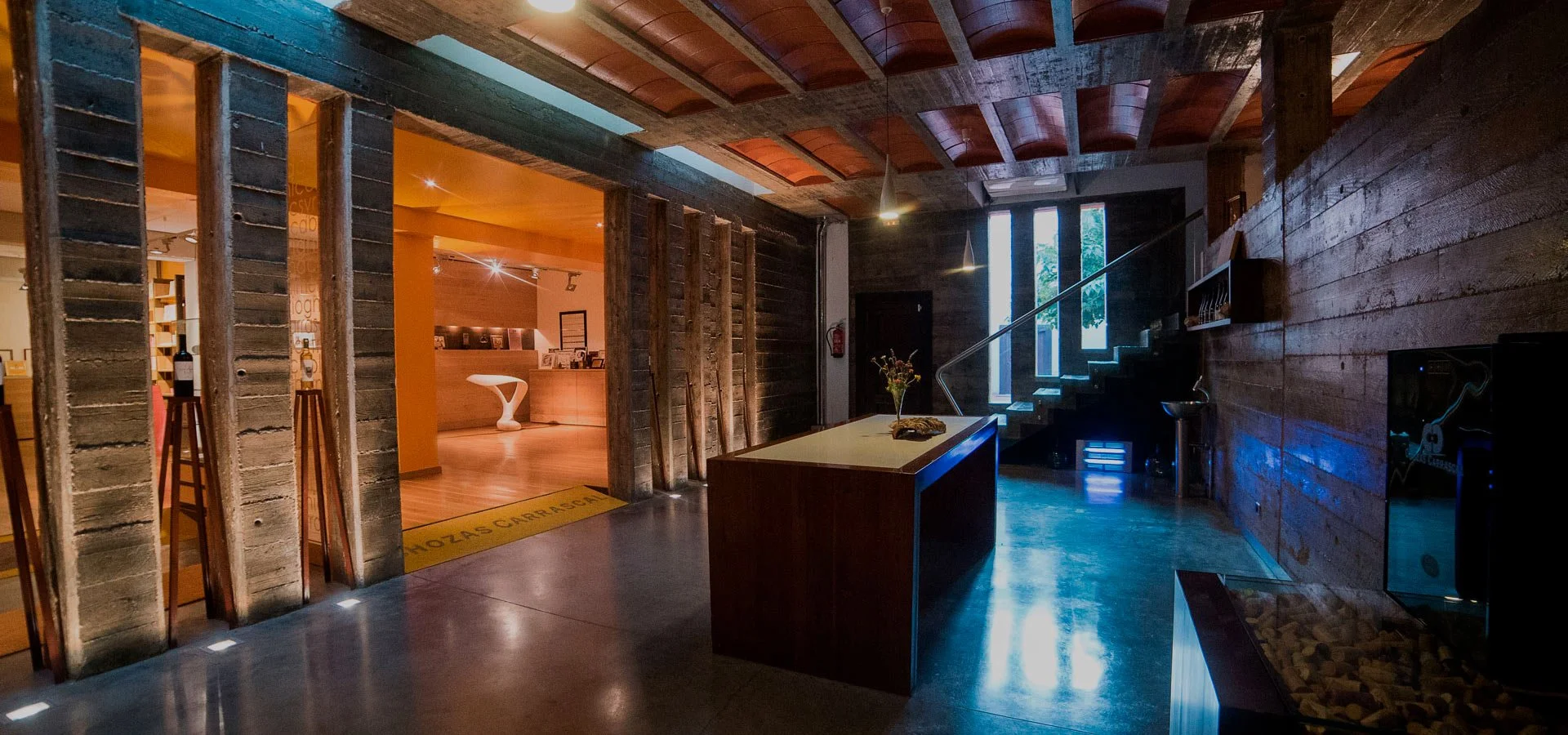 Interior of a modern building with a wood-paneled wall, concrete floor, staircase, and a reception desk. Soft lighting and natural light coming through tall windows.