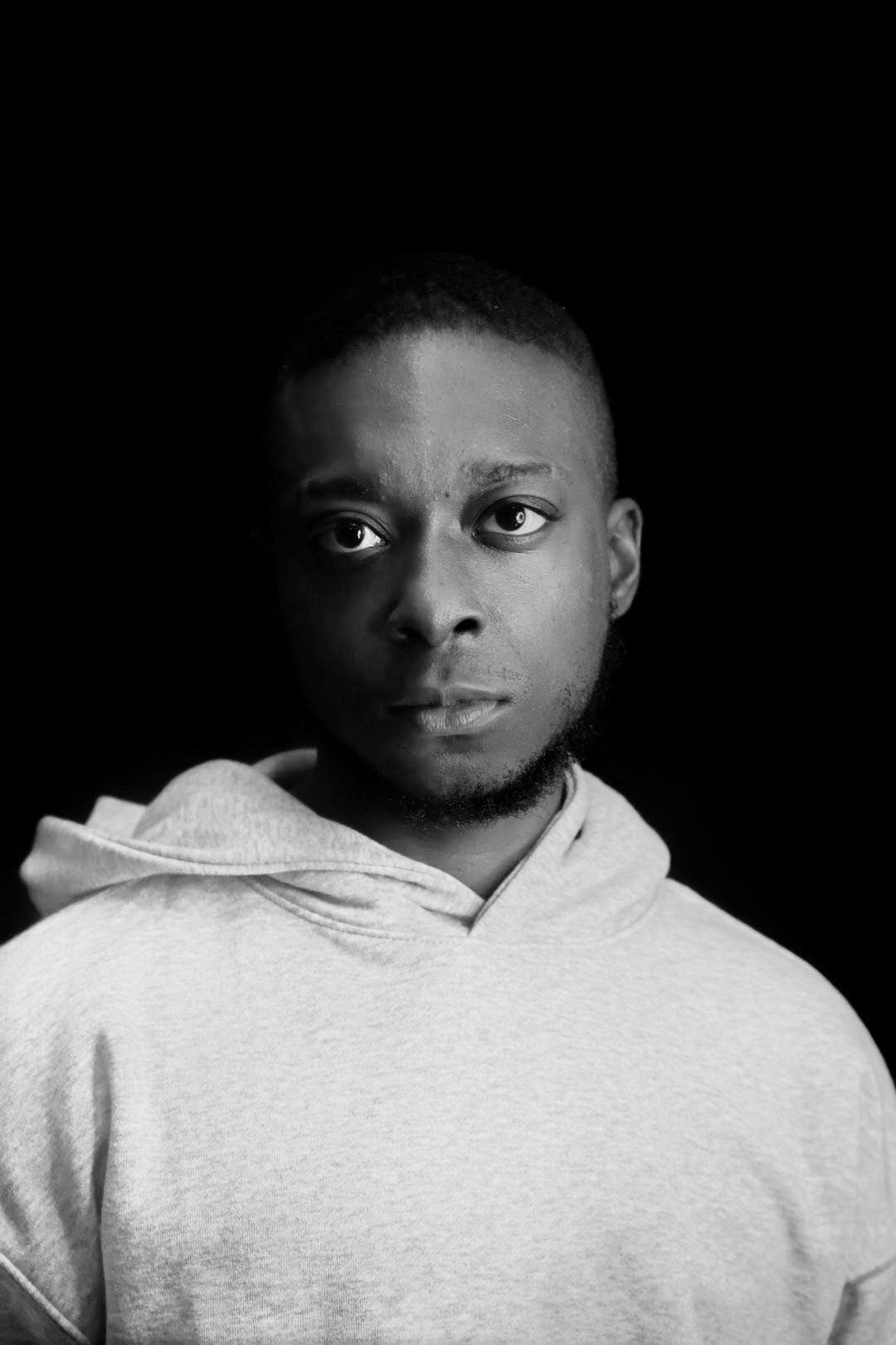 Black-and-white portrait of a young man with short hair, wearing a light-colored hoodie, against a dark background.
