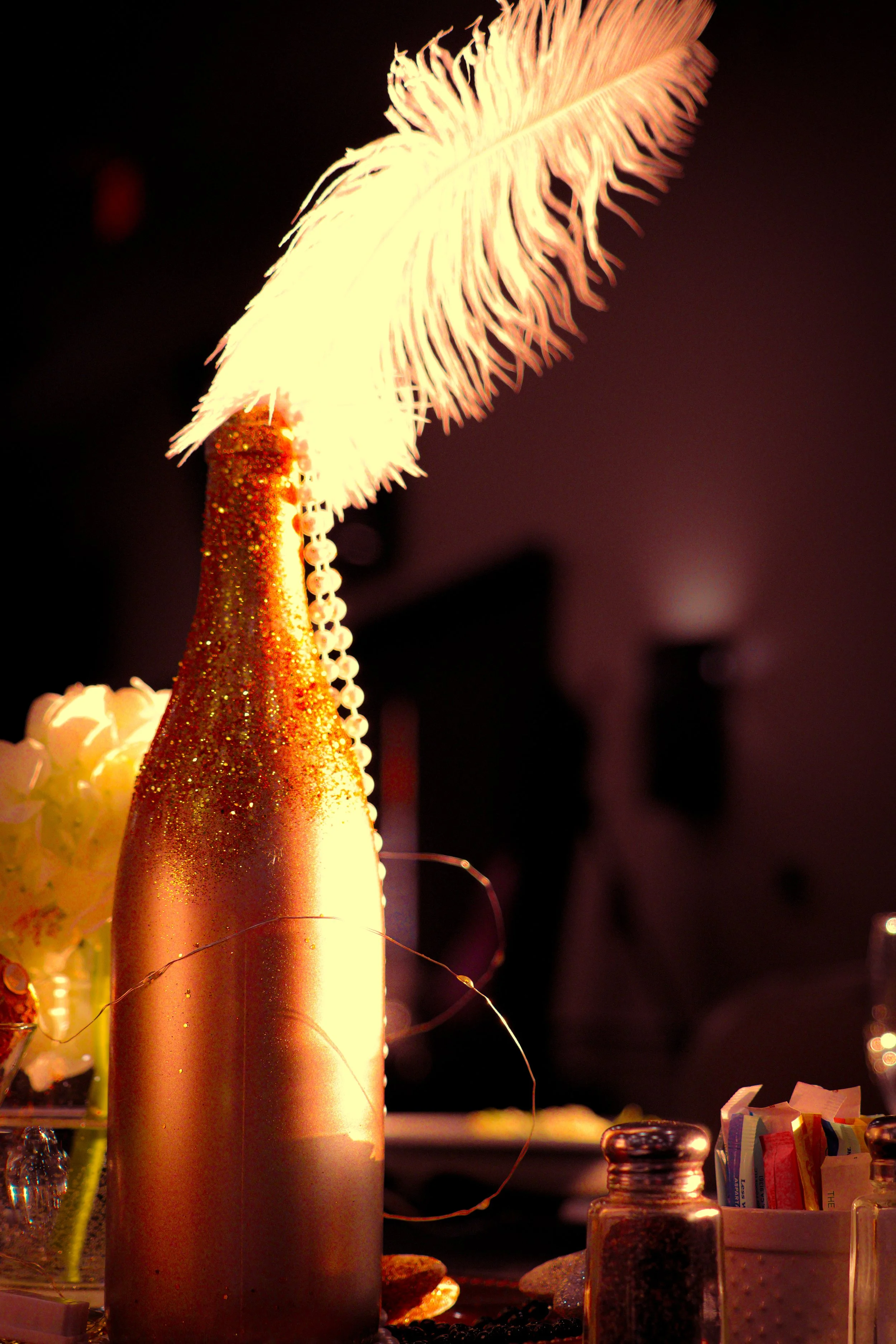A gold-tapered candle with a lit feather-shaped flame on top, surrounded by small objects and bottles on a dark background.