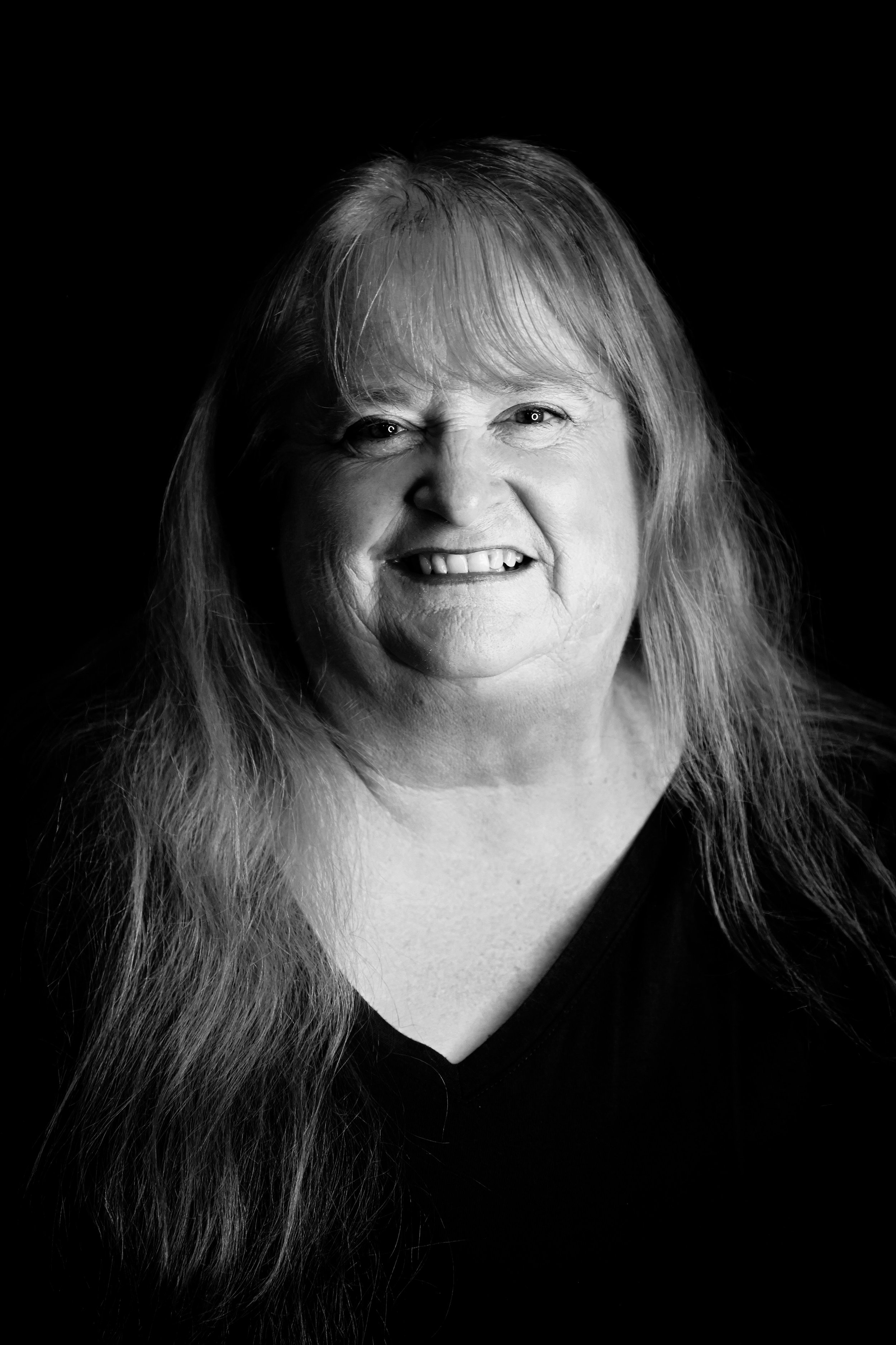 Black and white portrait of a smiling woman with long hair, parted in the middle, wearing a dark top against a plain dark background.