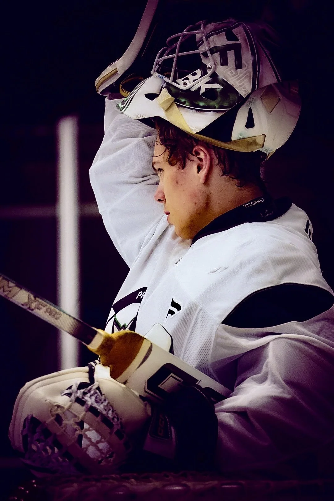 Young male hockey player wearing a white jersey, brown gloves, and a helmet with the face mask lifted off, holding his helmet with one hand and a hockey stick, appearing to be in thought or resting.