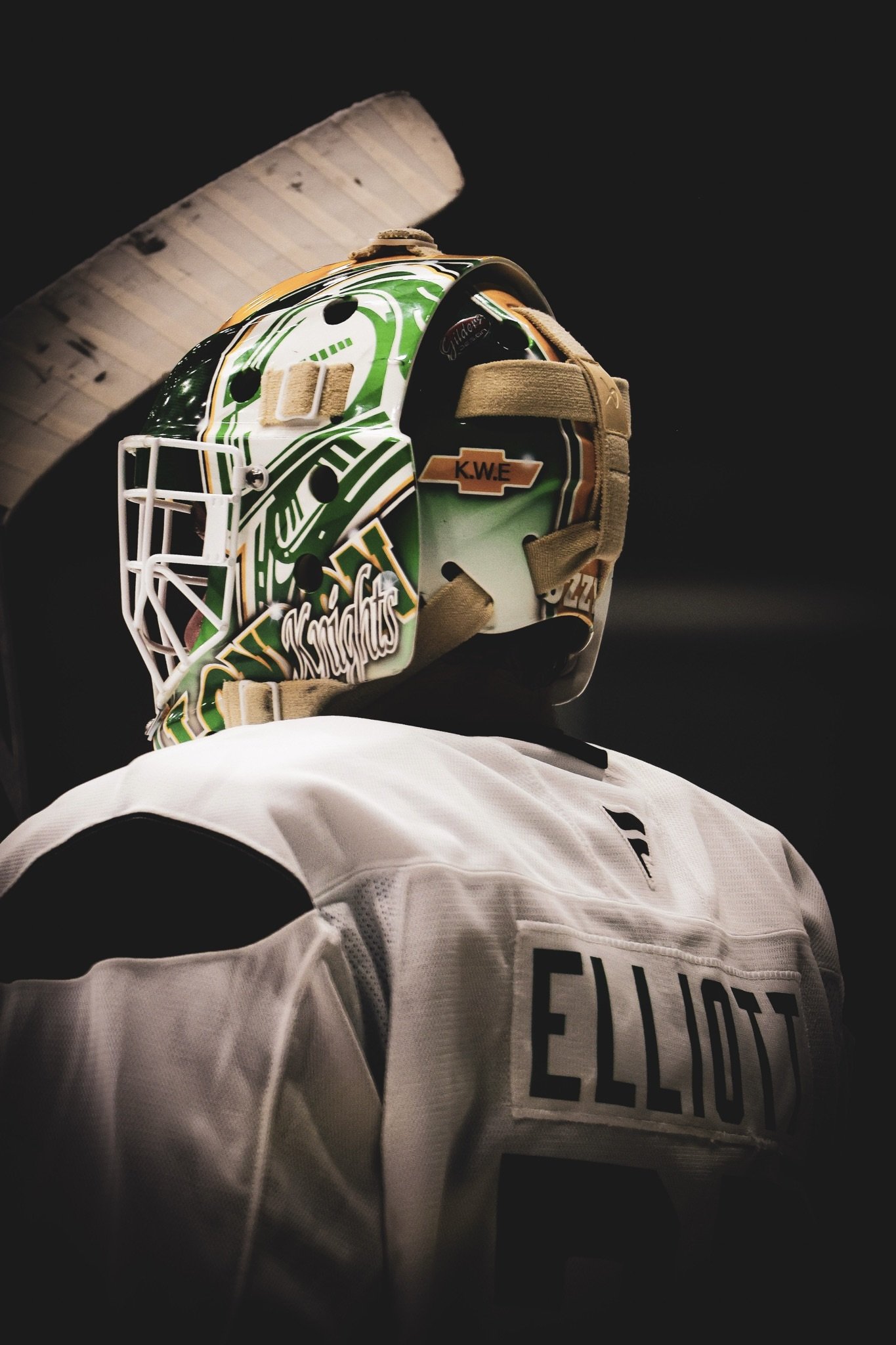 A hockey goalie helmet with a green, yellow, and white design featuring the words "Knights" and a black and white logo. The helmet is worn with beige straps. Part of a white hockey jersey with the name "Ellis" visible is also shown.