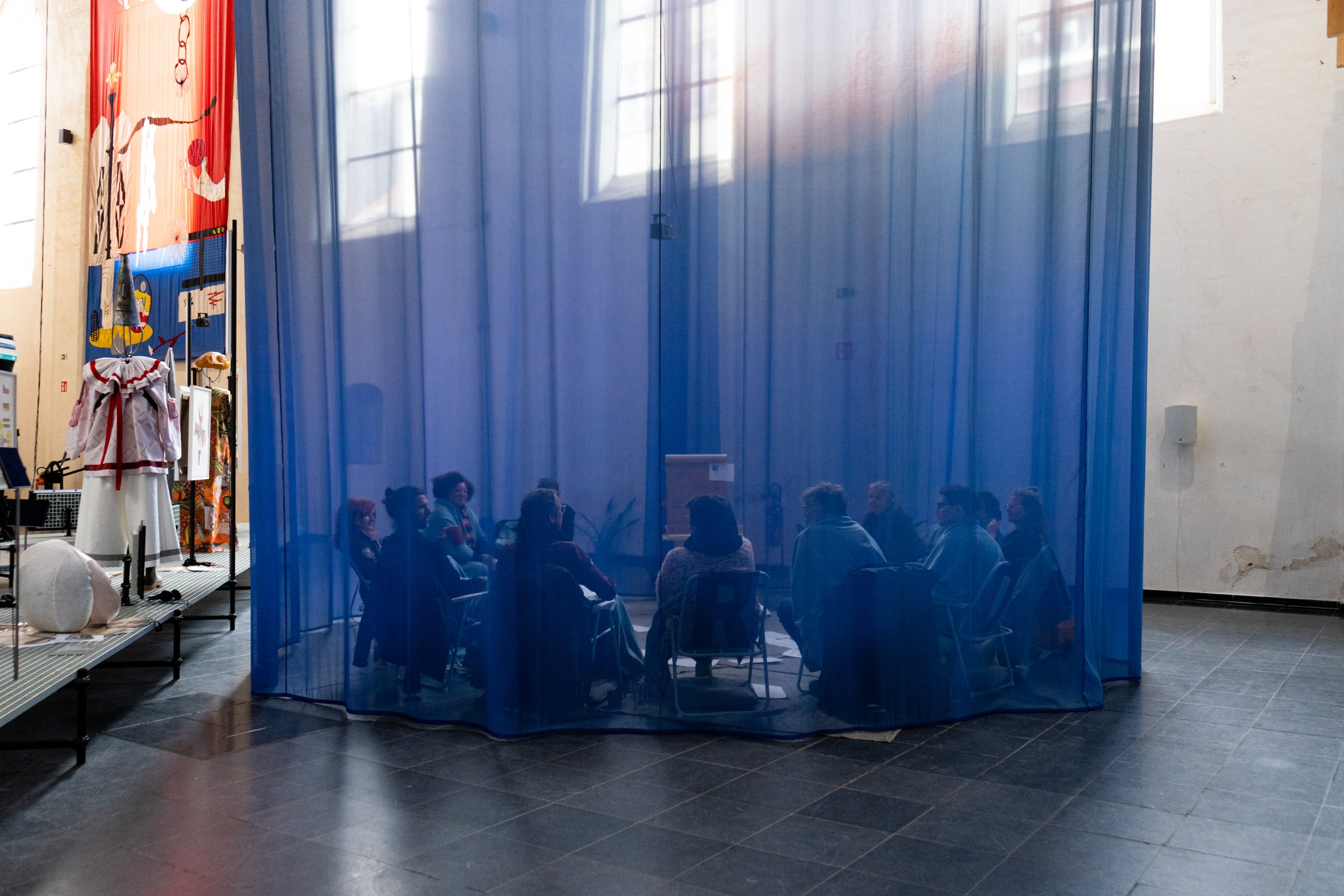 Groep mensen zit in een cirkel in een ruimte, afgezet door blauwe gordijnen, met ramen die natuurlijk licht binnenlaten.