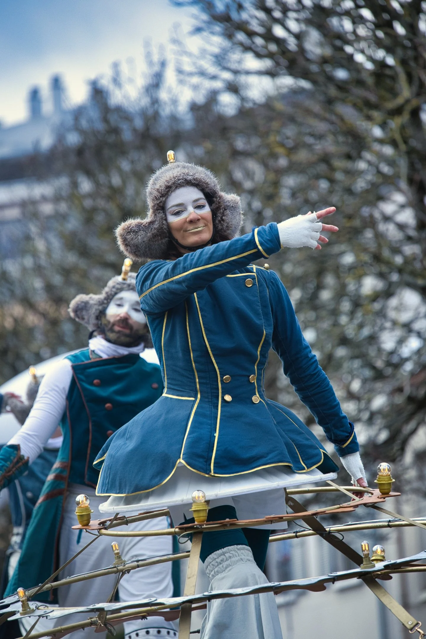 Des personnes déguisées en personnages de styles fantastiques dans un défilé. La personne en avant porte un costume bleu avec des détails dorés, un chapeau en fourrure, et se trouve sur une structure ressemblant à une plateforme circulaire avec des a