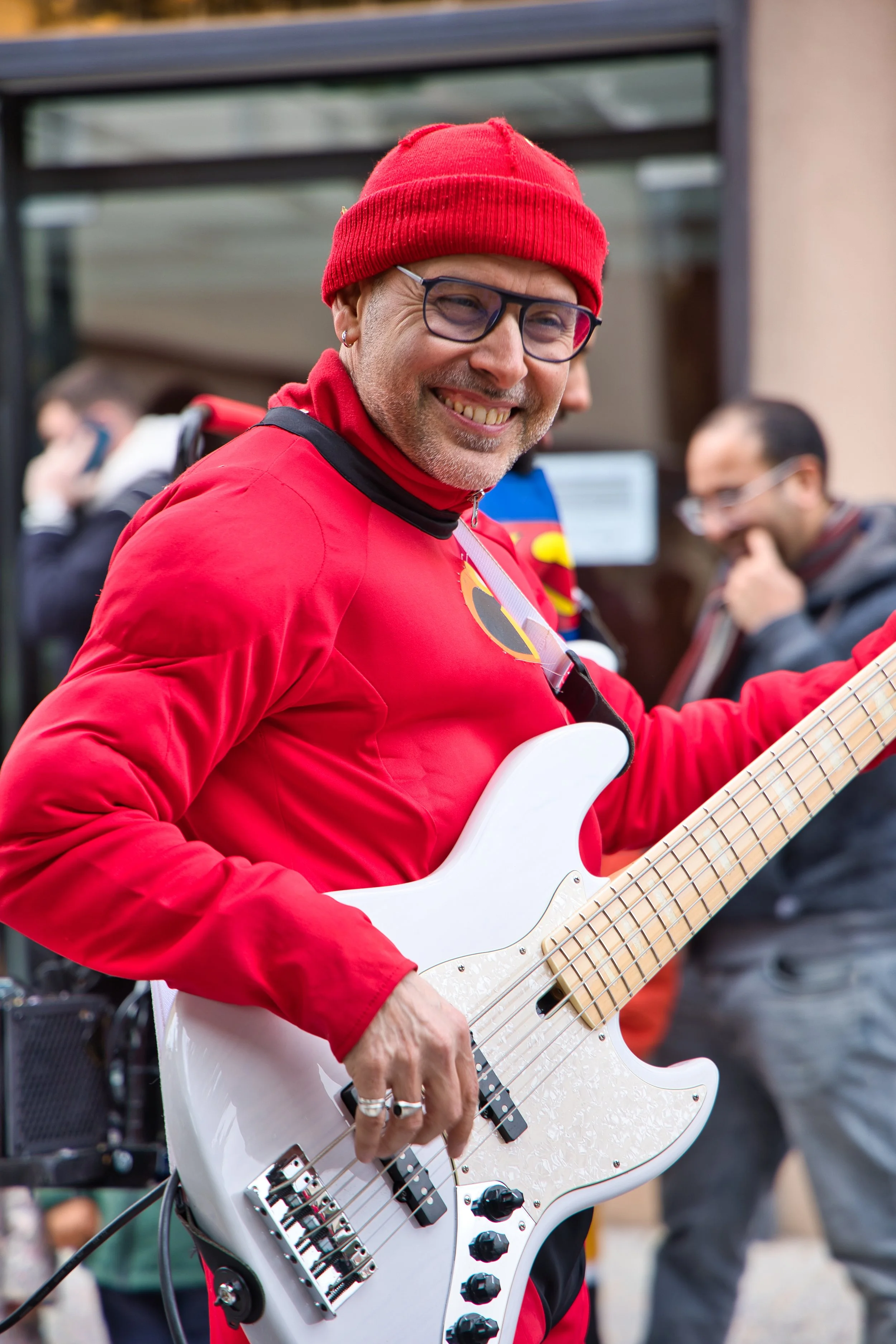 Un homme souriant portant un bonnet rouge, lunettes et un manteau rouge, jouant de la basse électrique blanche lors du carnaval de Thiais