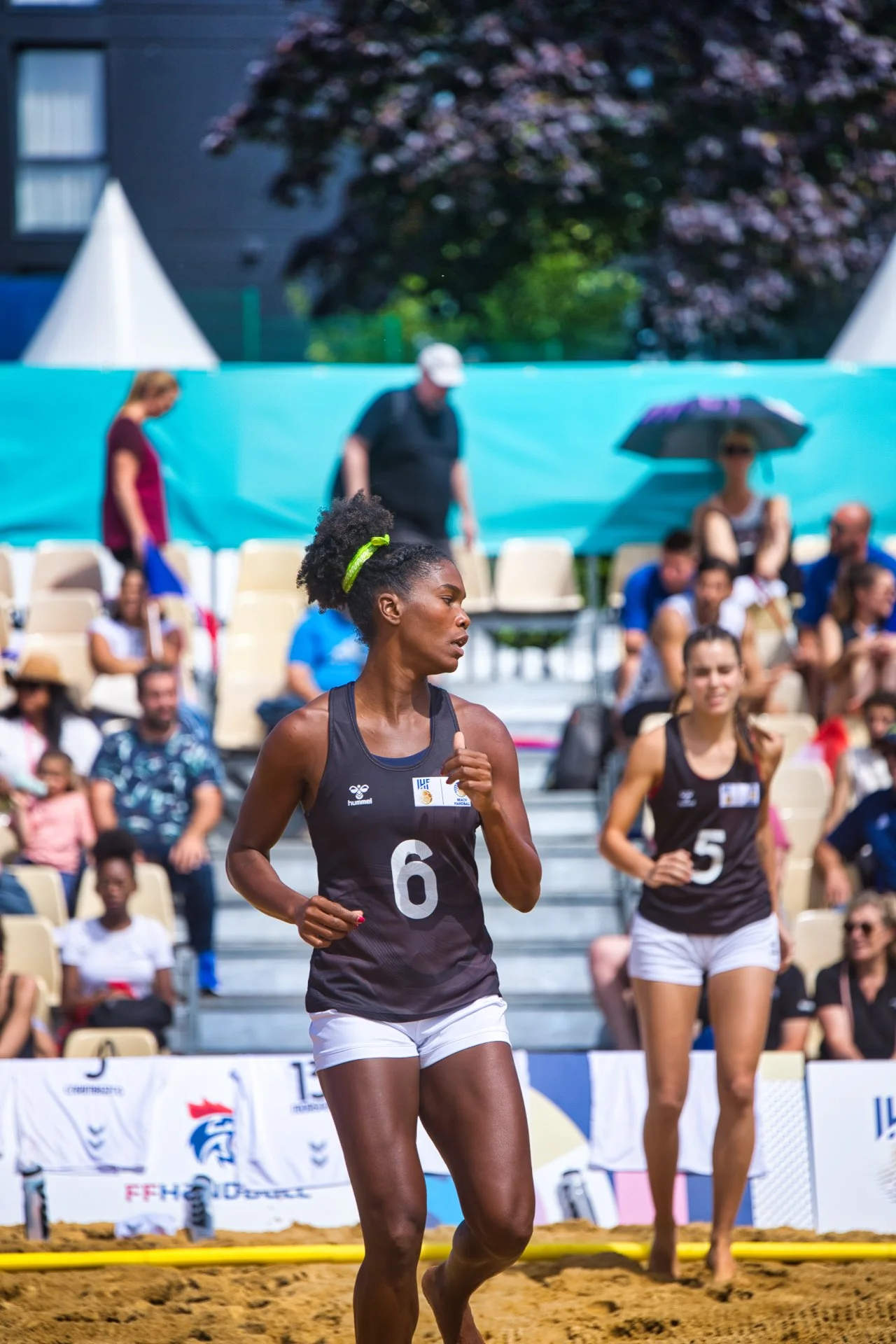Deux femmes jouent au beach-volley sur une plage, avec un public assis en arrière-plan et un ciel ensoleillé.