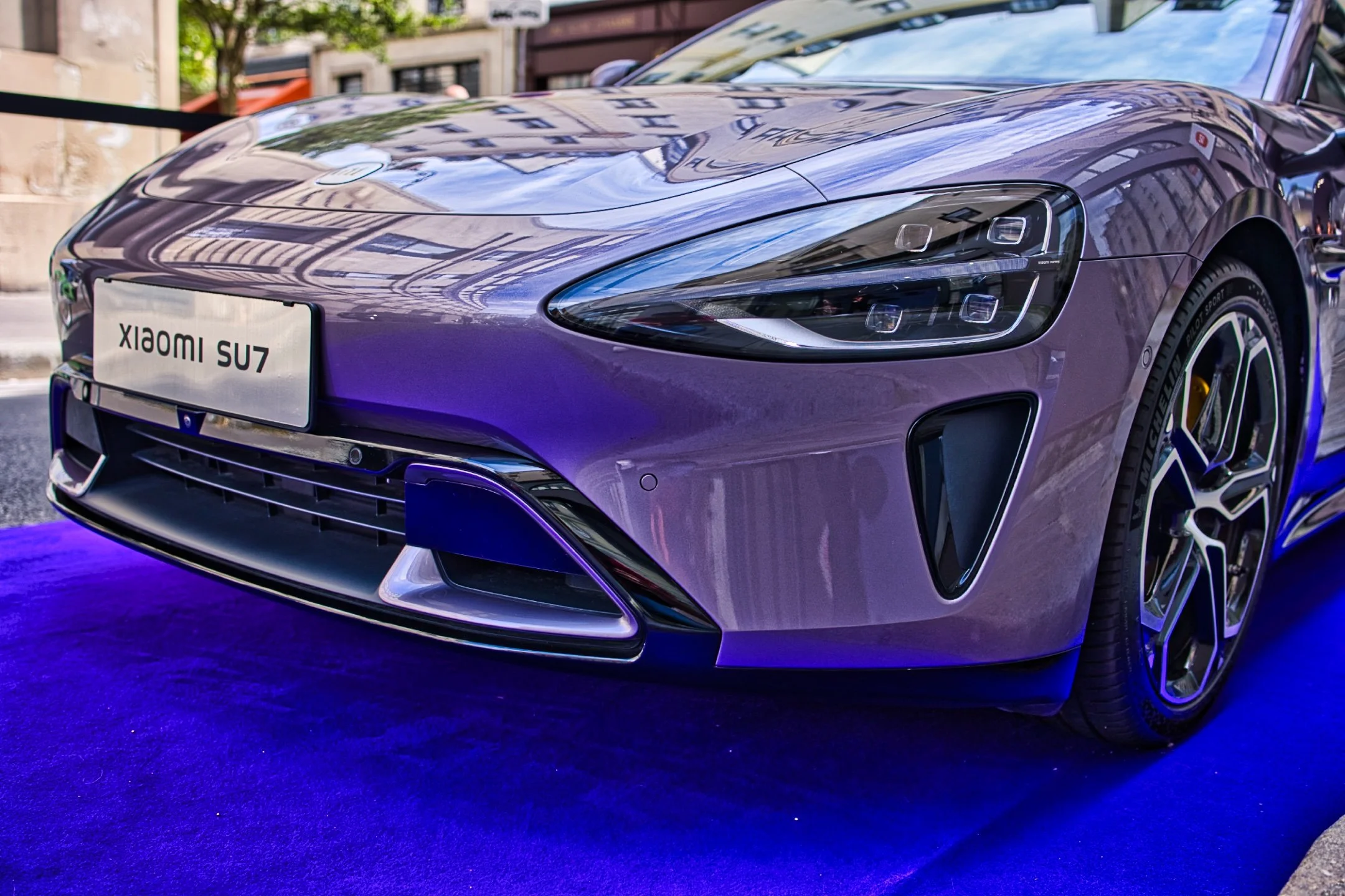 Xiaomi SU7 Max violette exposée sur un tapis bleu, avec un pare-brise reflétant des bâtiments urbains