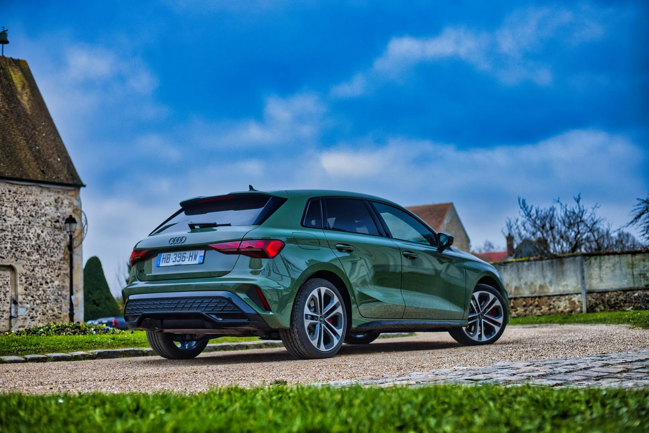 Audi A3 45 TFSIe verte stationnée sur une route en gravier devant des bâtiments anciens avec ciel bleu orageux.