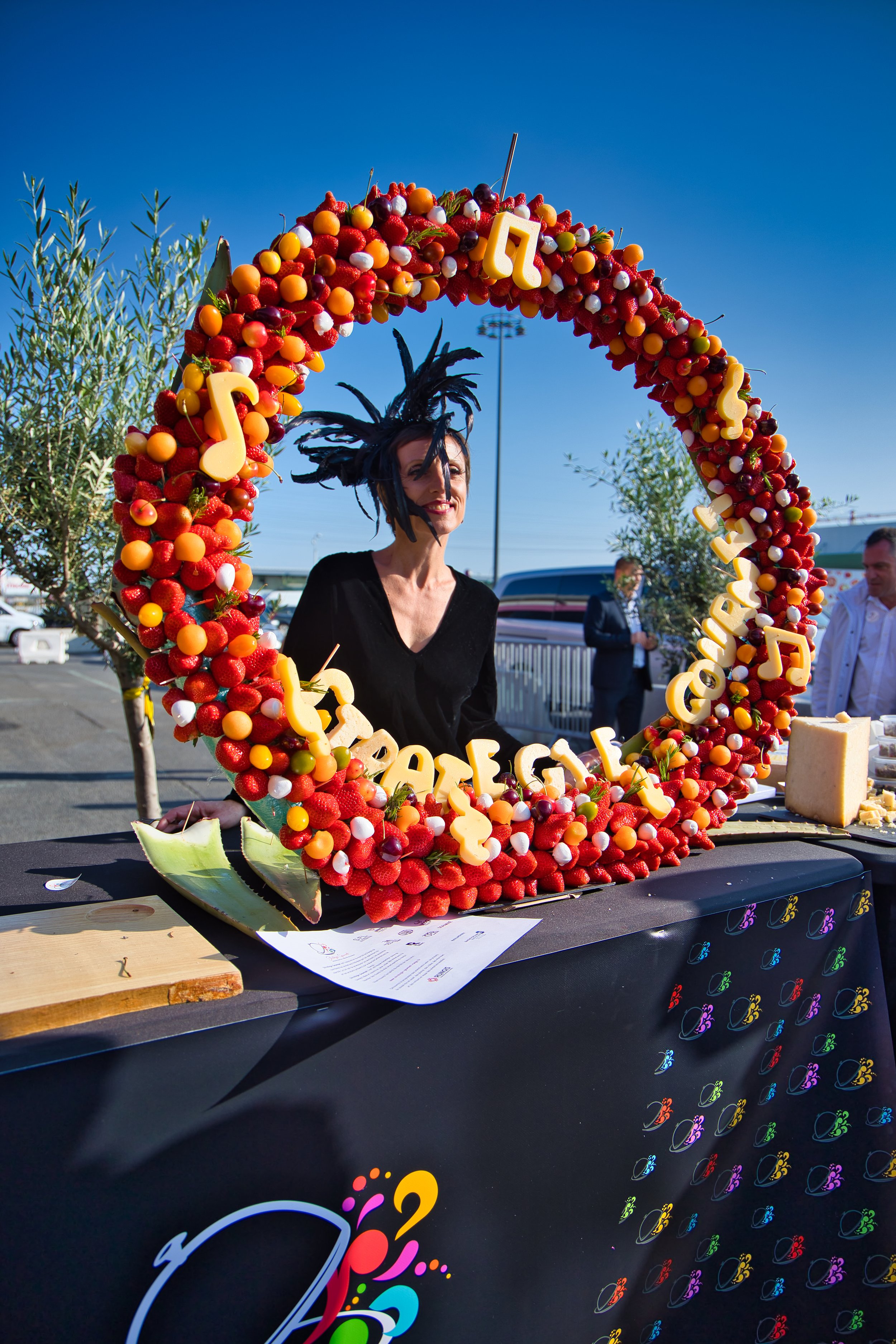 Une femme se tient derrière une table décorée d’un large cercle en fruits et bonbons, avec des lettres en fromage formant un message, lors d’un événement extérieur par temps ensoleillé.