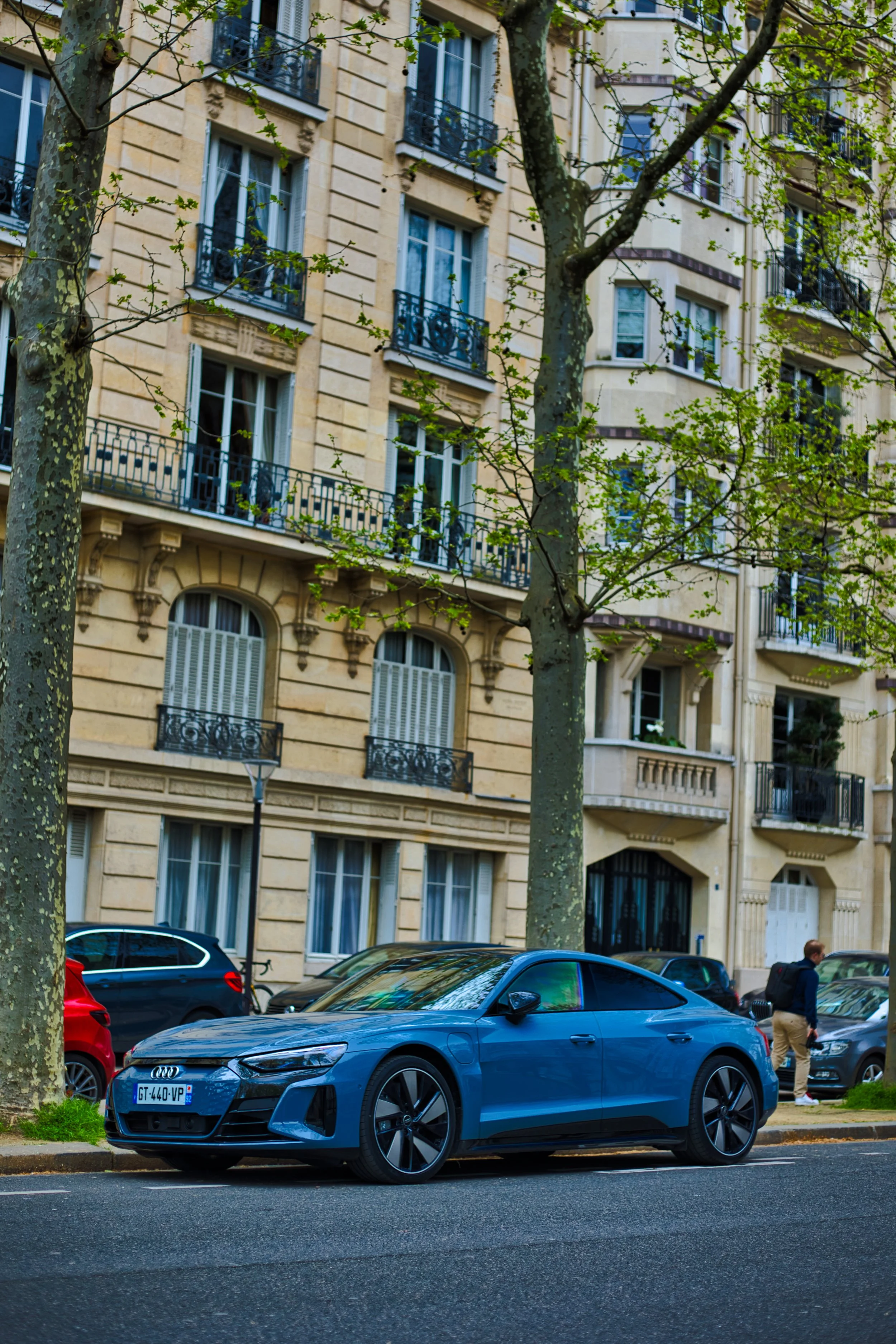 Une voiture bleue stationnée sur le côté d'une rue, avec un immeuble résidentiel en arrière-plan. Des arbres bordent la rue, une personne marche sur le trottoir.
