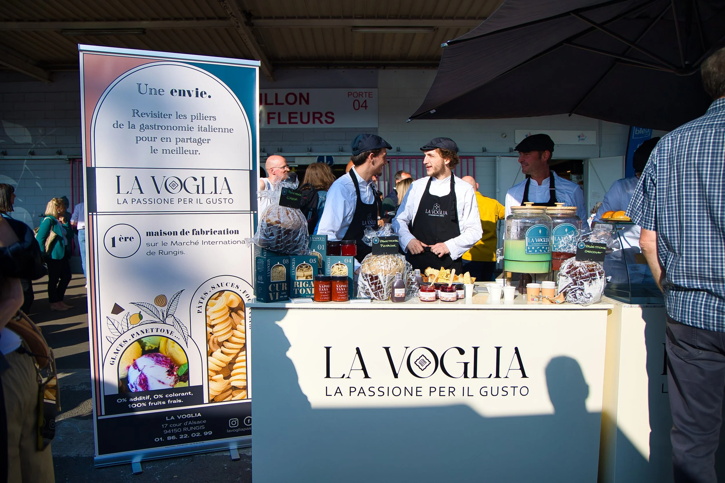 Stand de vente de produits italiens avec trois vendeurs en tenues noires et blanches, proposant des confitures, jus et biscuits. À gauche, une grande pancarte promotionnelle avec texte en français et italien. Le stand porte le nom 'LA VOGLIA'.