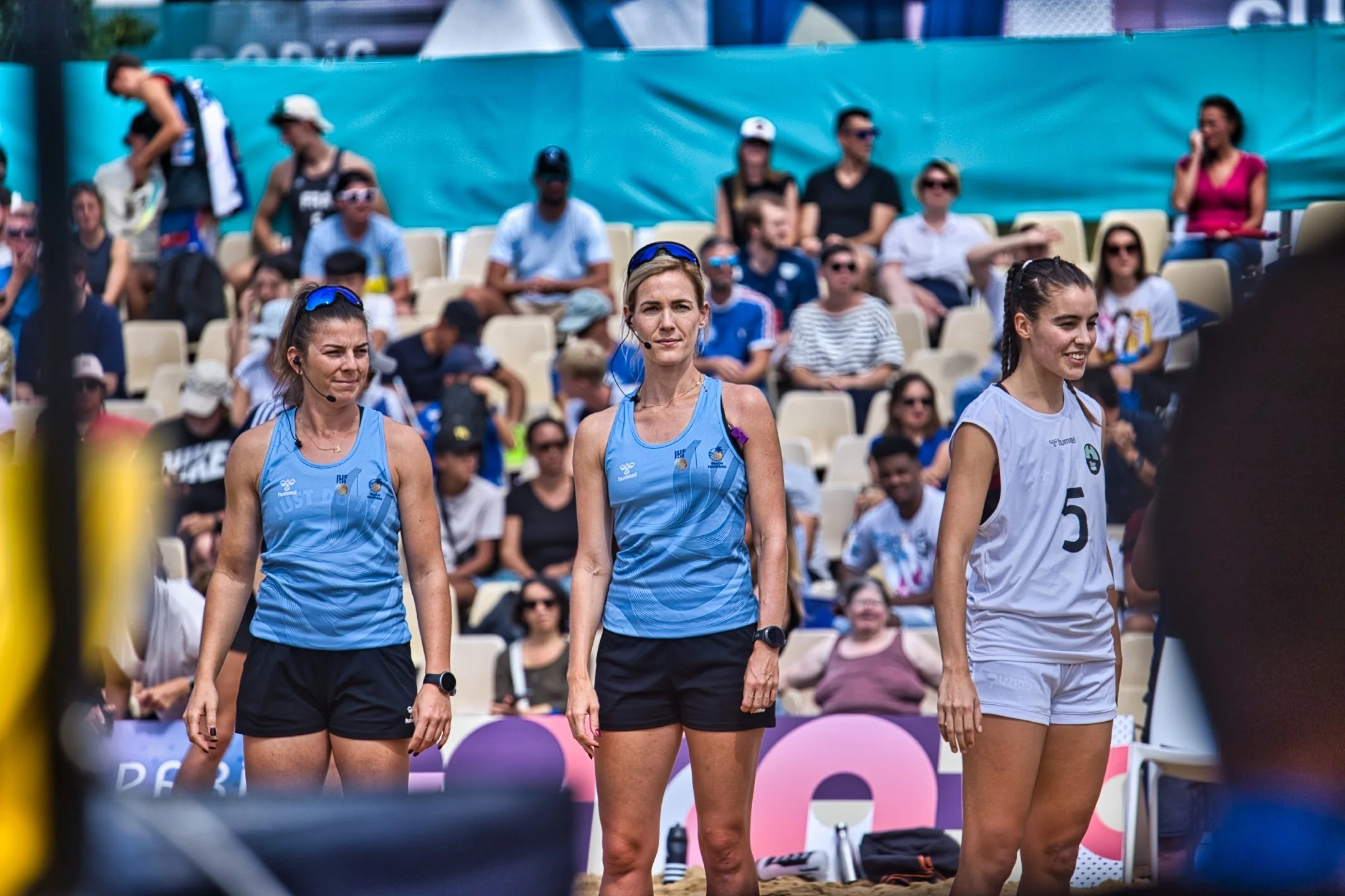Trois joueuses de beach handball en tenue sportive, deux en bleu et une en blanc, lors d'une compétition, avec un public assis en arrière-plan, lors des Jeux de Paris 2024