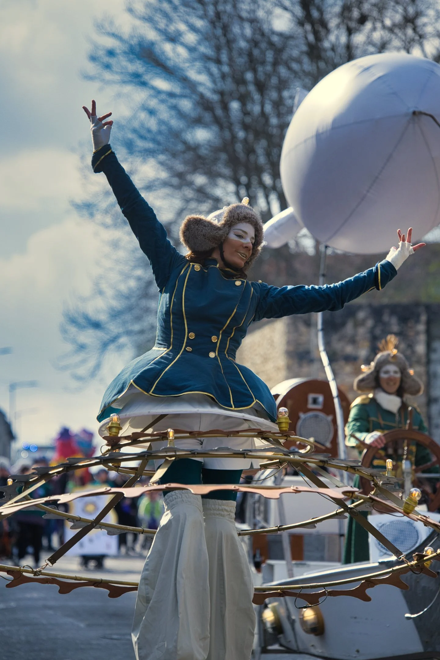Fête avec personnes en costumes victoriens, dont une femme sur une structure mécanique avec bras levés, fond d’un ciel partiellement nuageux et des arbres dénudés lors d'un carnaval