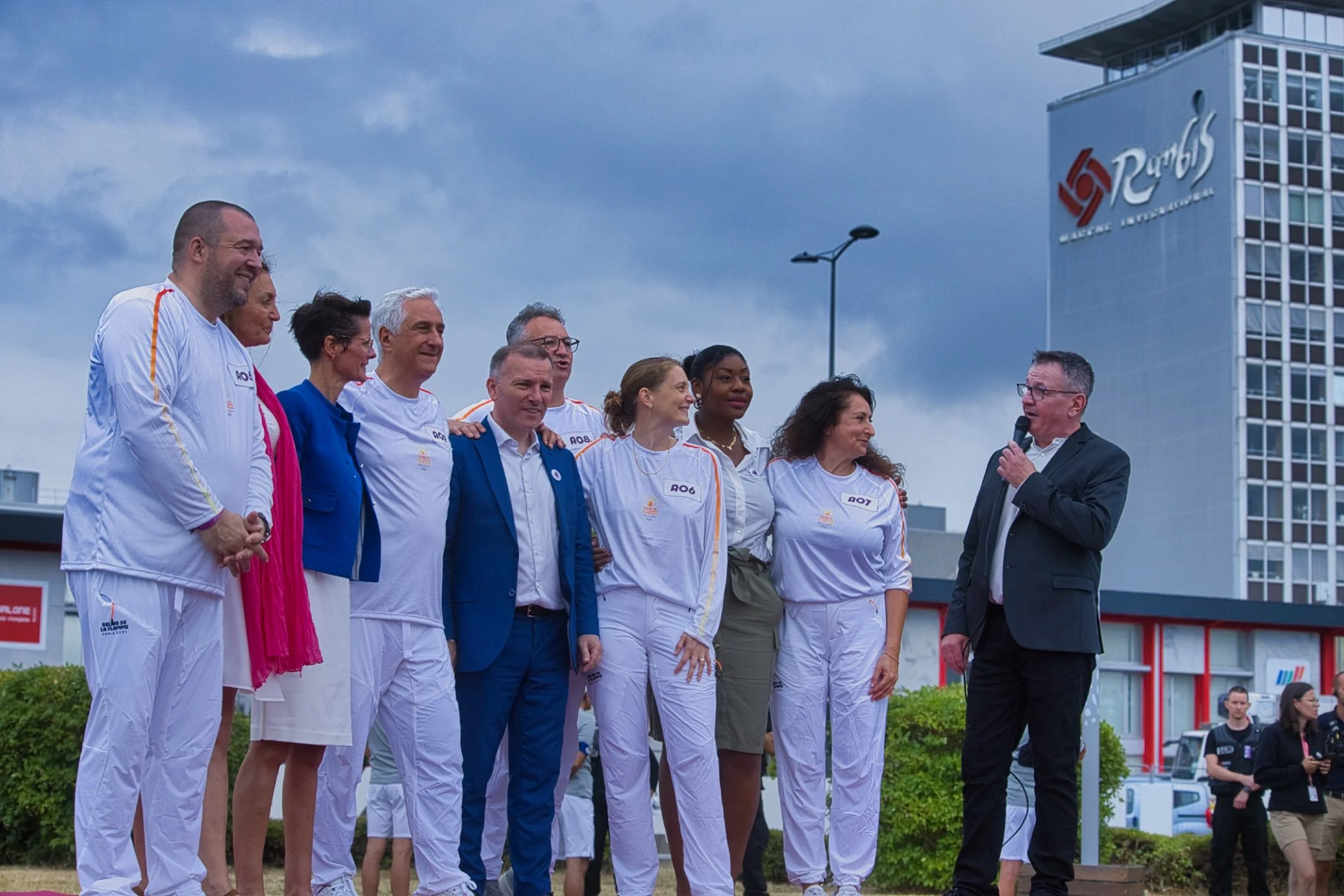Personnes en tenue blanche avec des accents orange, se tenant en ligne lors de la cérémonie de la Flamme Olympique JO Paris 2024, alors qu'un homme en costume noir parle dans un microphone, avec un bâtiment avec le logo ''Rungis en arrière-plan.