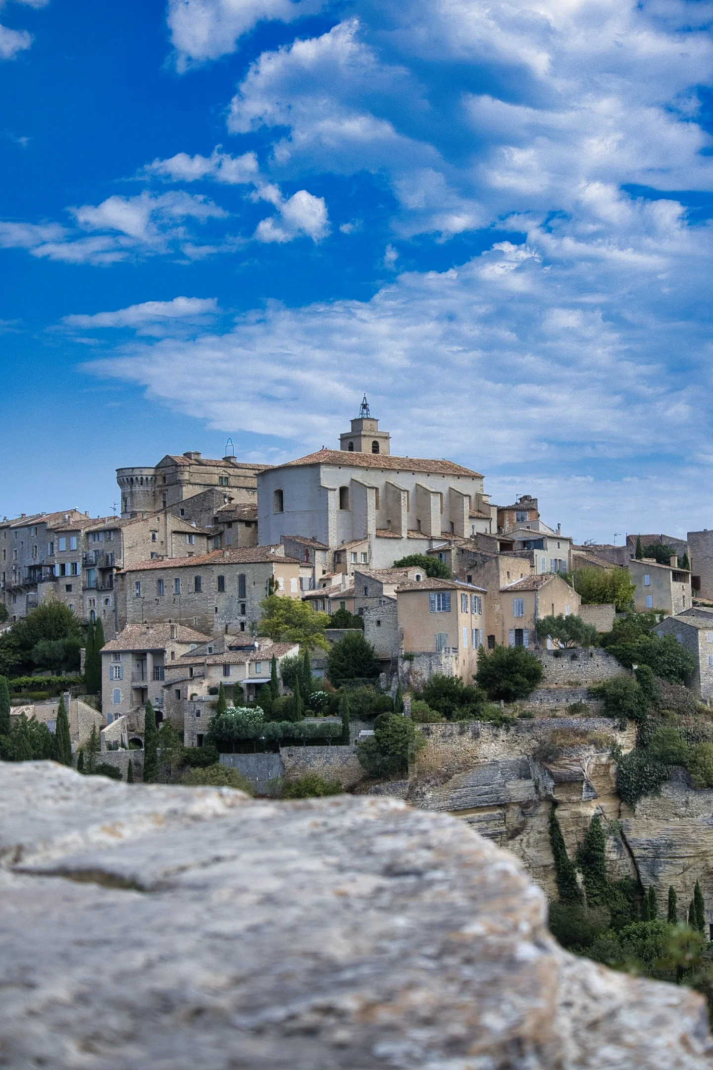 Vue d'un village médiéval en hauteur avec des bâtiments en pierre, un ciel bleu et quelques nuages.