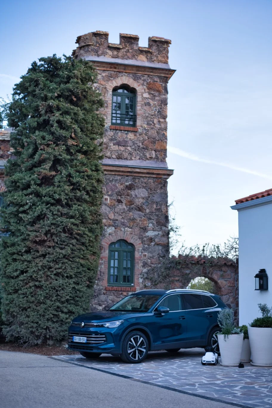 Volkswagen Tiguan noire stationnée devant un vieux bâtiment en pierre avec deux fenêtres à volets verts et un vieux arbre, sous un ciel clair.