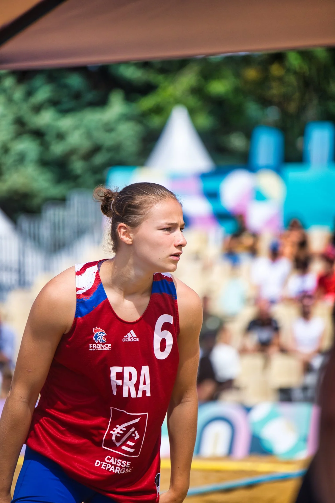 Joueuse de beach-volley en maillot rouge avec le numéro 6, tissu de France, lors d'une compétition en extérieur avec spectateurs en arrière-plan.