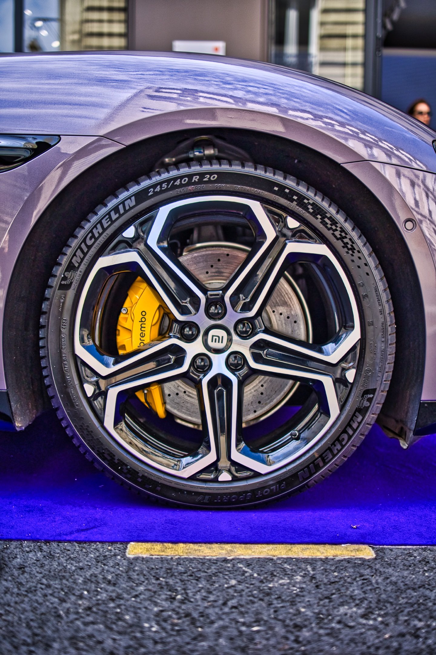 Roues d'une voiture électrique, avec des jantes noires et des freins Brembo jaunes, posée sur un tapis violet. Xiaomi SU7 Max