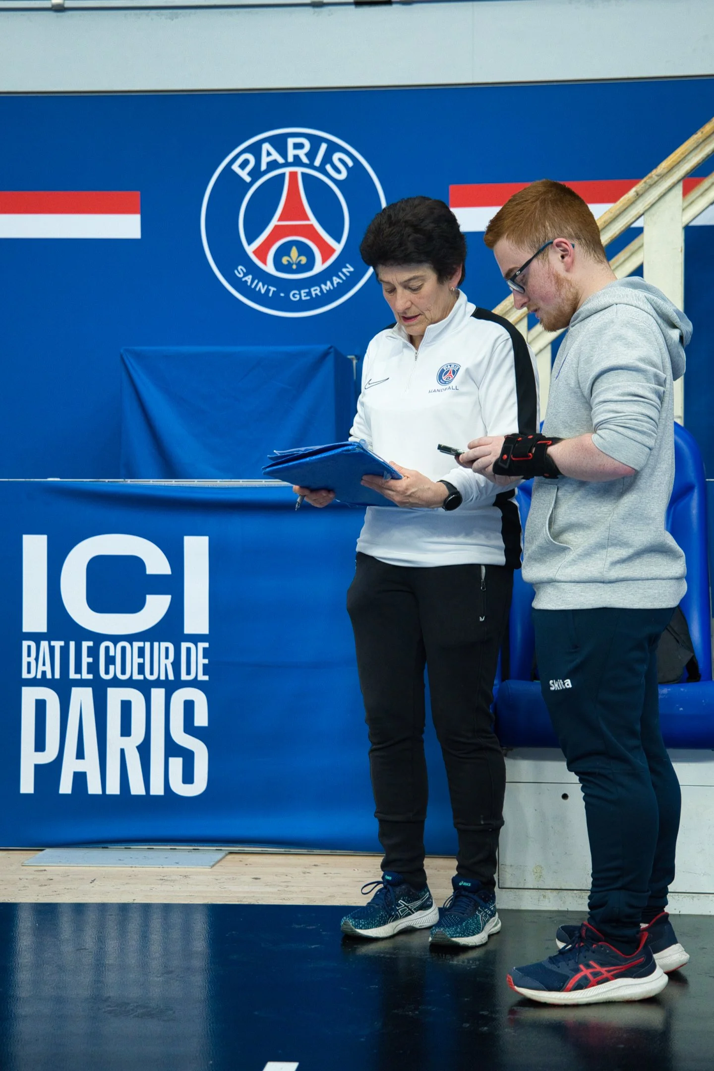 Deux personnes dans une salle d'entraînement sportif vérifient des documents, avec un mur en arrière-plan affichant le logo du Paris Saint-Germain.