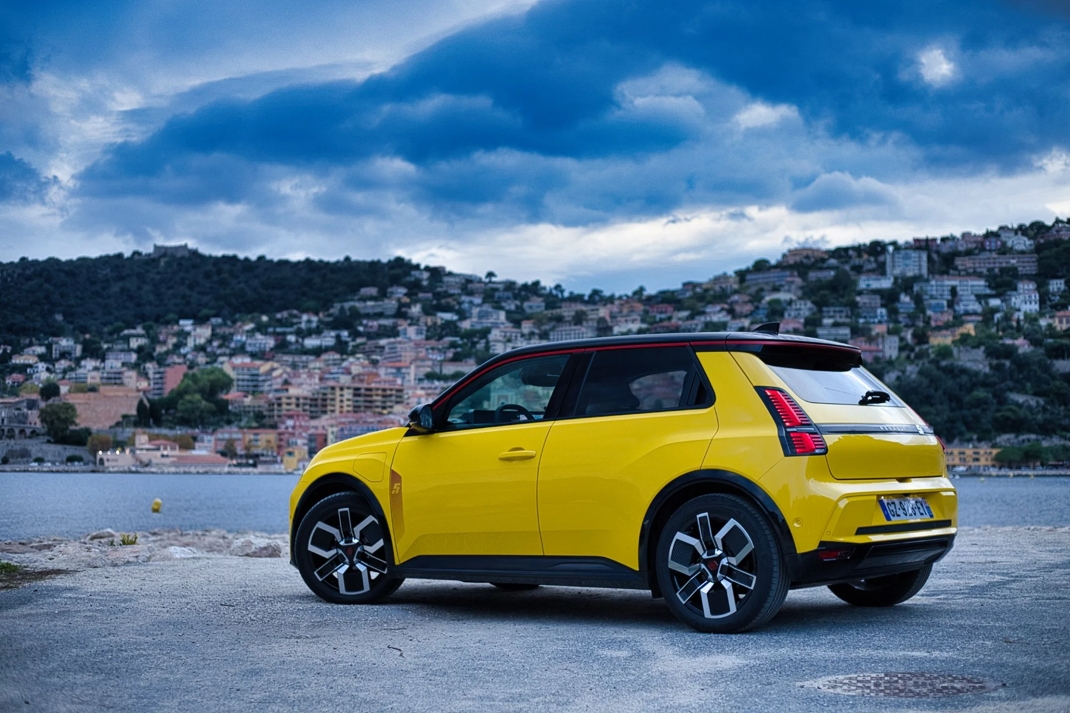 Renault R5 e-tech jaune stationnée près d'un lac avec une ville et des montagnes en arrière-plan, ciel nuageux.