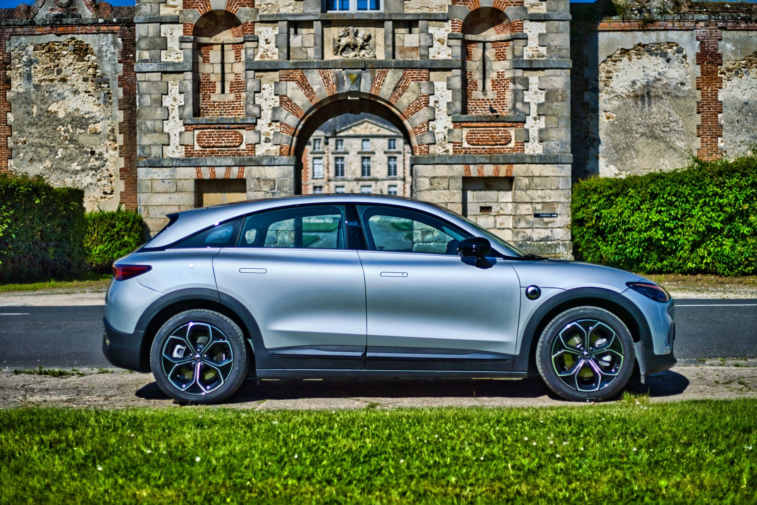 Smart #3 grise stationnée sur le bord de la route devant un mur en pierre antique avec un arc et un bâtiment visible par l'arche, entourée de végétation verte.