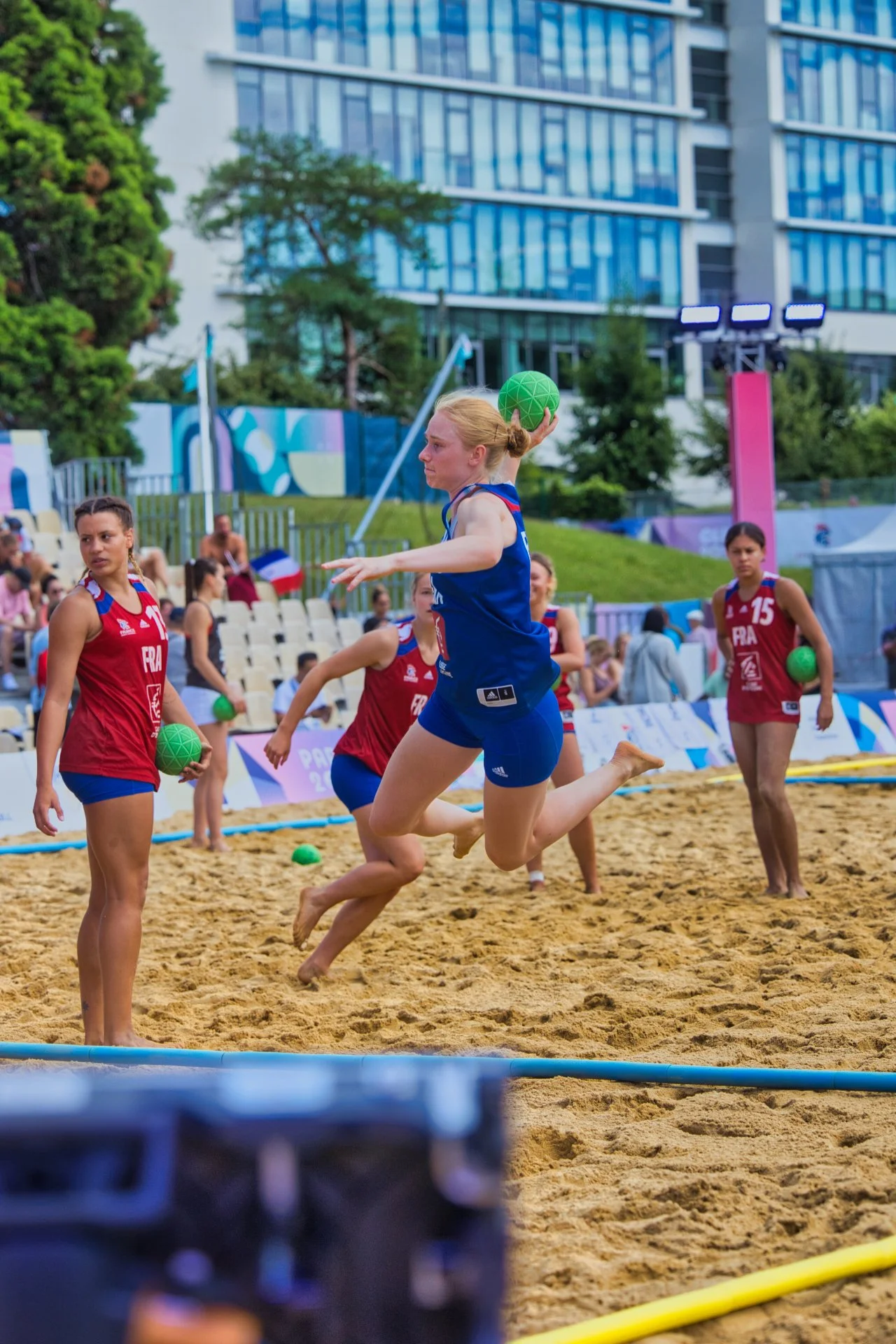 Des joueuses françaises de beach handball évoluent sur un terrain de sable, portant des maillots rouges et bleus, avec des ballons verts, en pleine action lors d'une compétition en extérieur, entourées de spectateurs et dans un cadre urbain. JO Paris