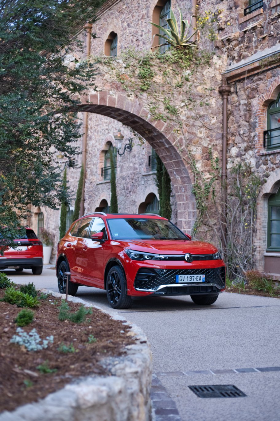 Volkswagen Tiguan rouge dans une cour pavée entourée de bâtiments en pierre avec des arches et des fenêtres vertes, avec de la végétation environnante.