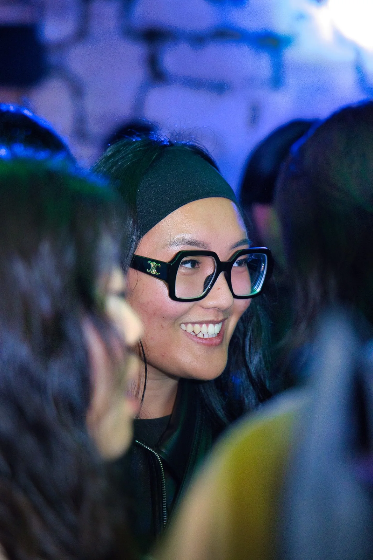 Une jeune femme souriante portant des lunettes épaisses et un bandeau noir, entourée de plusieurs personnes.