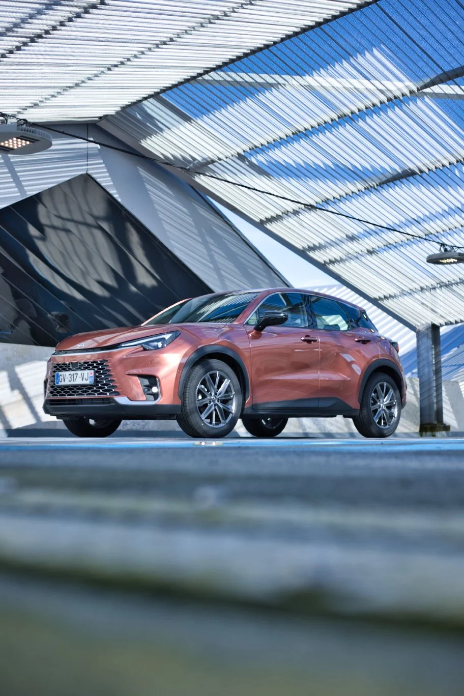 Lexus LBX de couleur bronze garée sous une structure moderne en métal avec des lignes en diagonale, dans un environnement urbain contemporain.