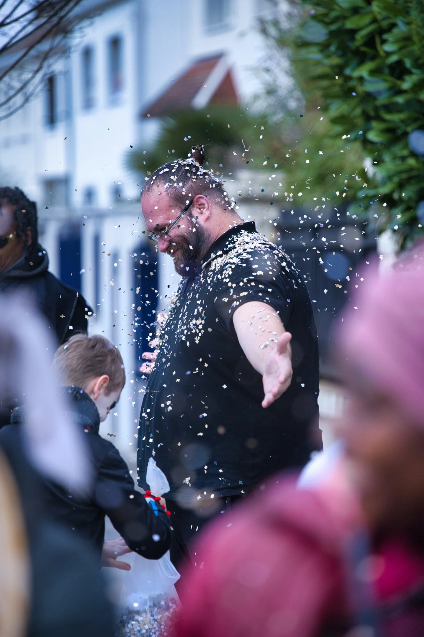 Un homme avec des lunettes et une barbe, souriant, entouré de confettis, lors d'une célébration en extérieur. Plusieurs autres personnes sont également présentes, dont un enfant qui joue avec le confetti lors d'un carnaval