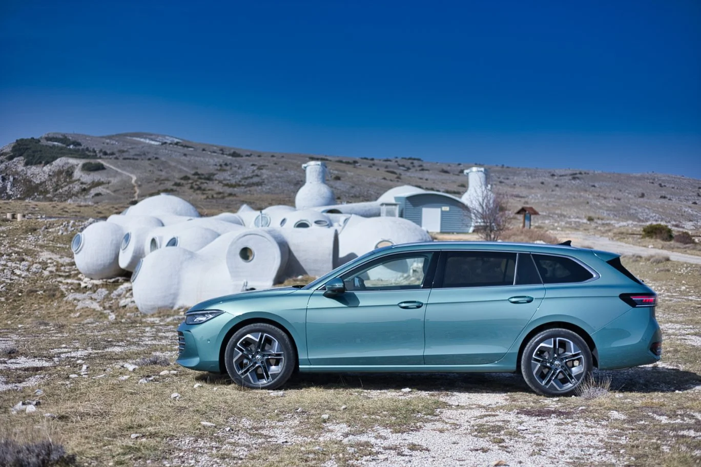 Volkswagen Passat de couleur verticoptere stationnée sur un terrain rocheux avec une architecture futuriste blanche en forme de capsules devant un paysage de collines sous un ciel bleu clair.