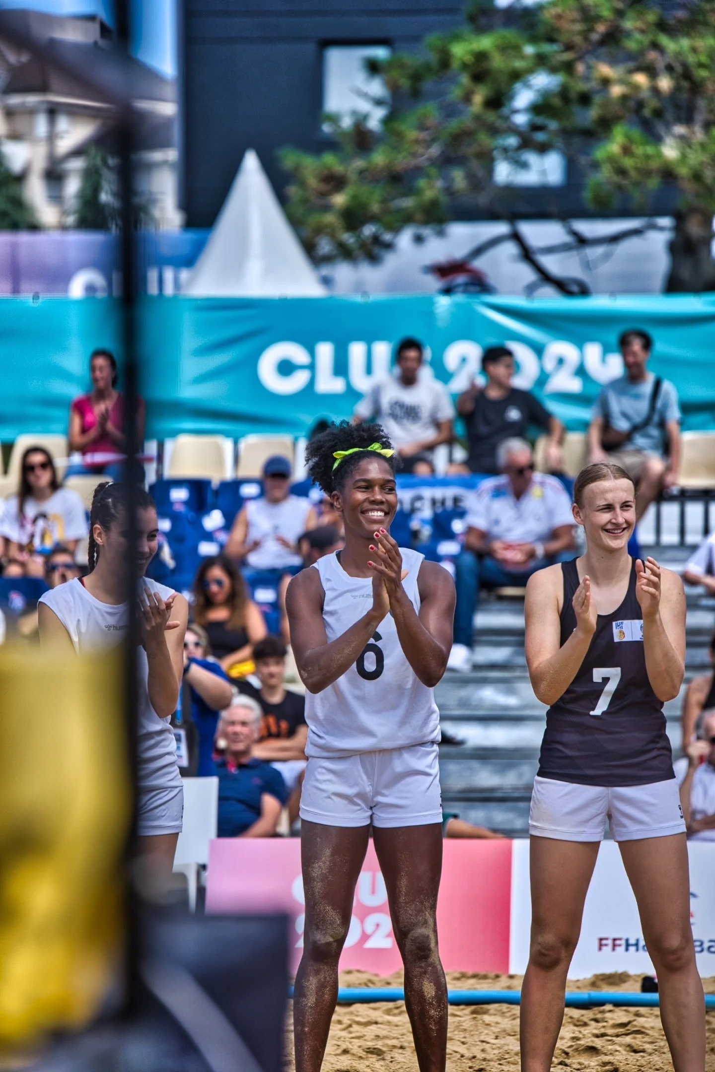 Trois joueuses de beach handball en tenue de sport, applaudissant et souriant, lors d'un match ou d'une cérémonie, avec des spectateurs en arrière-plan et un panneau indiquant 'CLUB 2024' lors des Jeux de Paris 2024