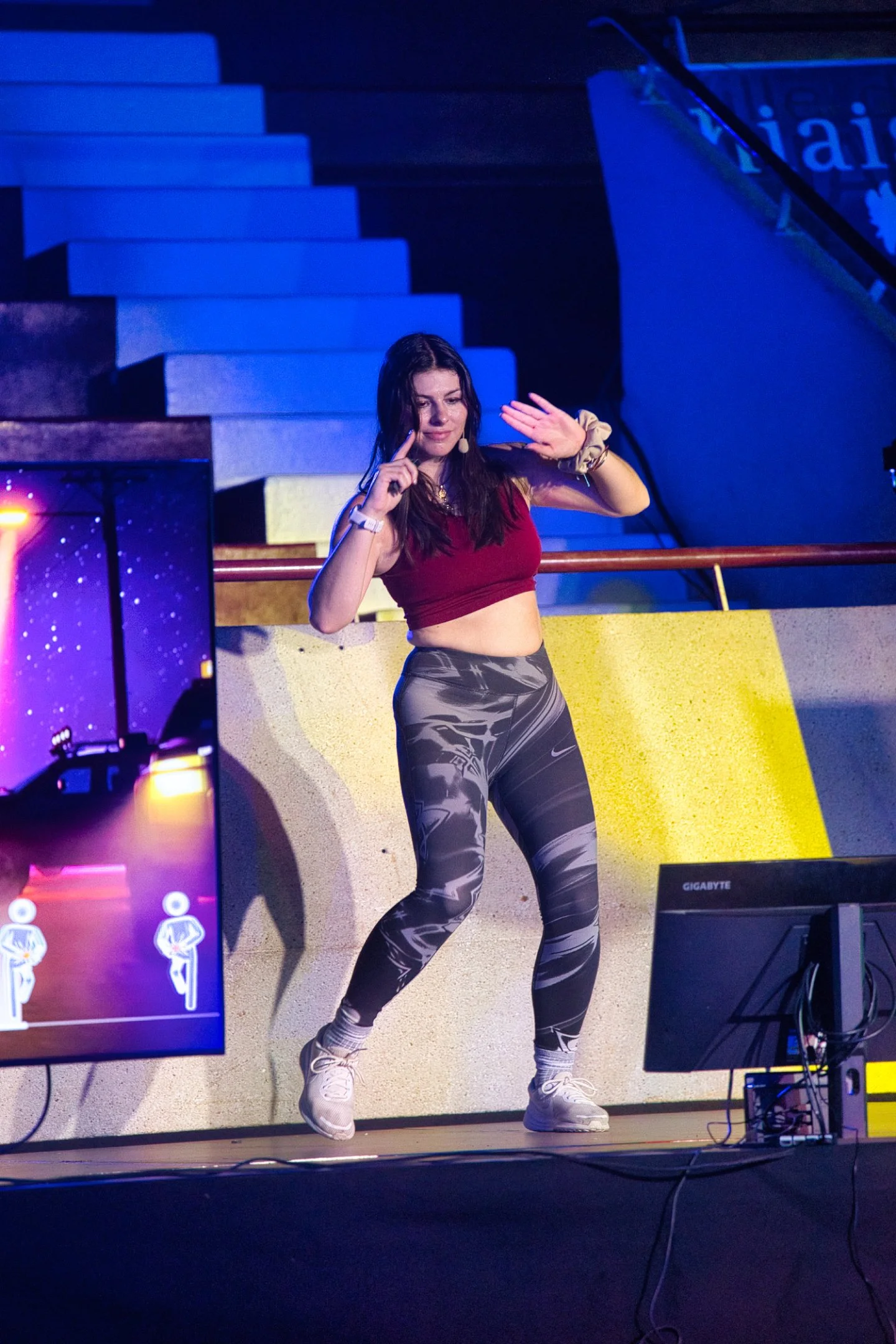 Une jeune femme danse sur scène, portant un haut rouge et un legging noir et blanc, avec des lumières colorées en arrière-plan lors de l'évènement Gaming Théodasium 2025 à Thiais