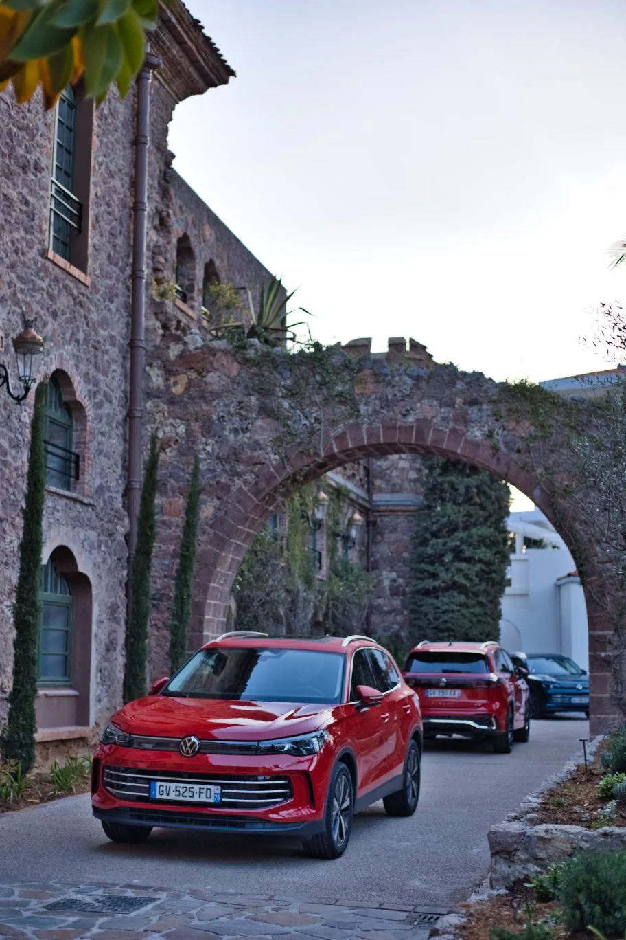 Volkswagen Tiguan rouges garées dans une rue pavée devant une vieille bâtisse en pierre avec une arche en pierre sur le côté, entourée de végétation.