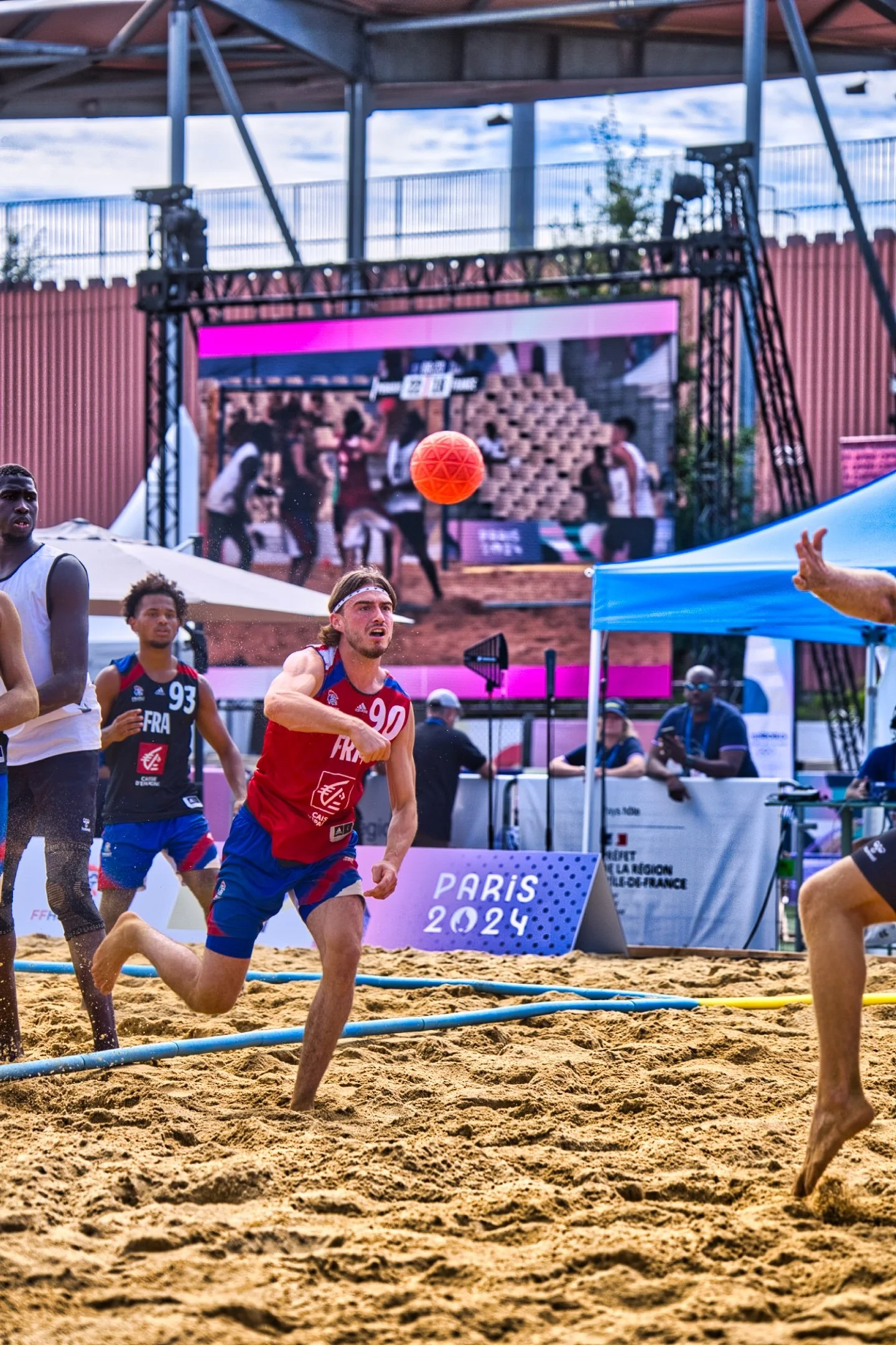 Des joueurs de beach handball en pleine action lors des Jeux de Paris 2024, avec un écran géant en arrière-plan affichant la même scène.