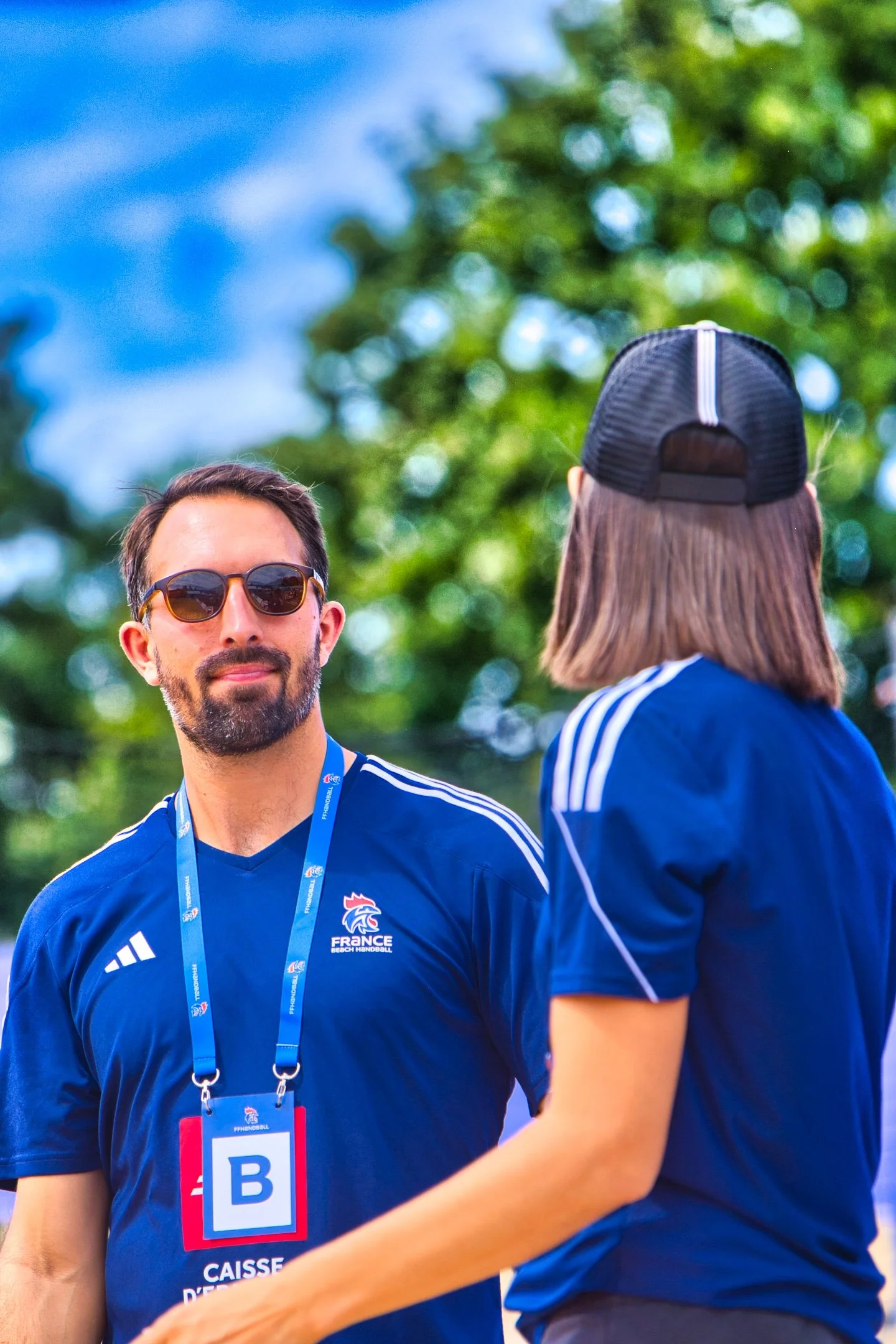 Deux personnes portant des maillots de l'équipe de France de handball discutent dehors, avec des arbres en arrière-plan. lors des Jeux de Paris 2024