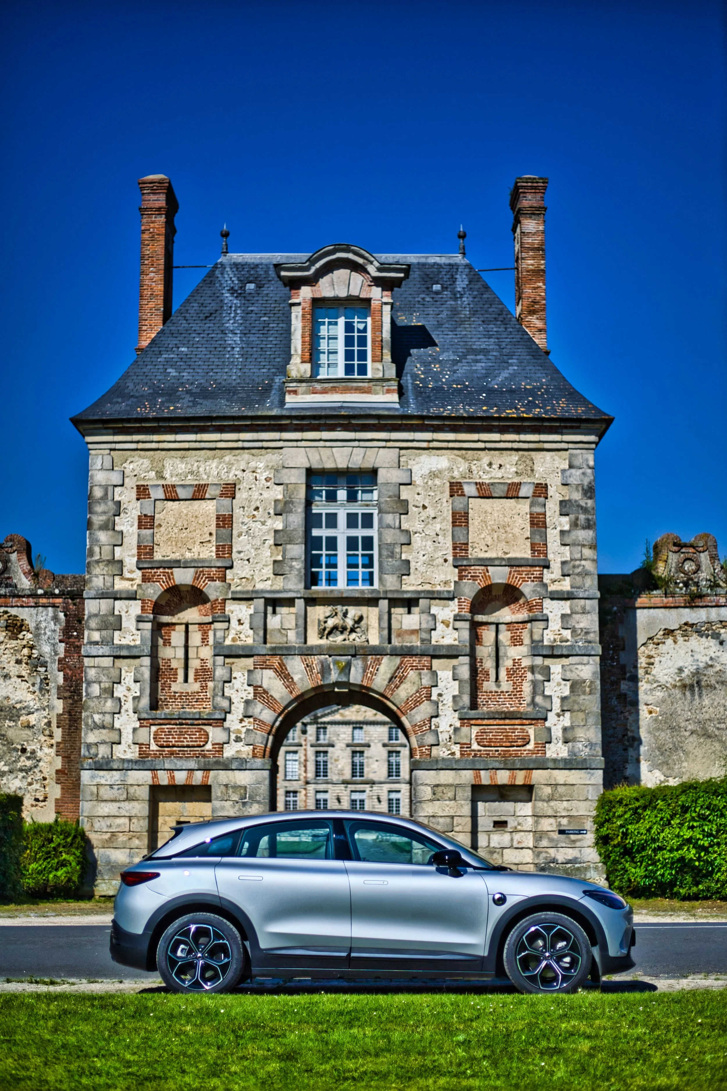 Smart #3 de couleur argentée garée devant un bâtiment ancien en pierre avec des murs en brique et des fenêtres à meneaux, sous un ciel bleu clair.