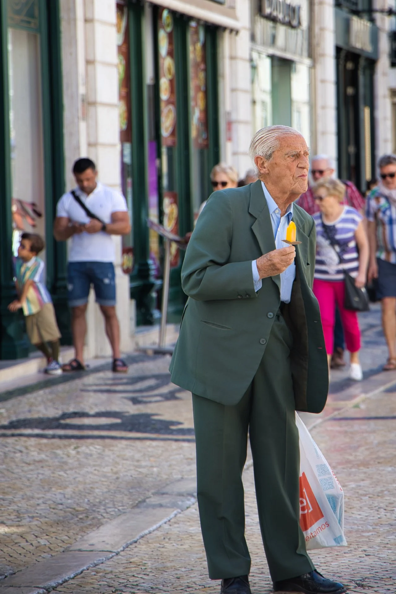 Un homme âgé en costume gris tient une bougie allumée dans une main, dans une rue animée avec plusieurs personnes en arrière-plan.