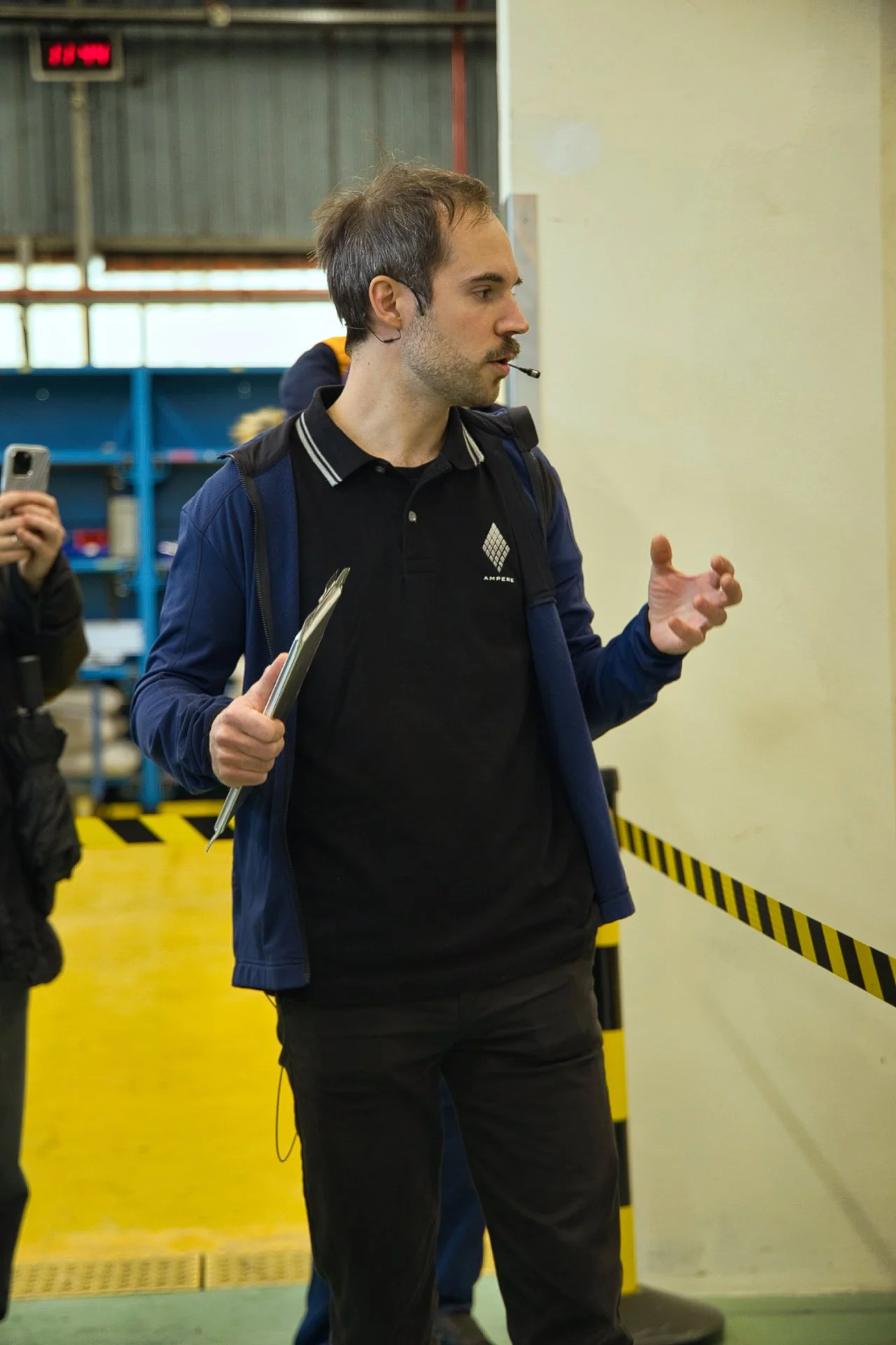 Un homme parle en direction d'une audience dans une salle industrielle, portant un casque sur l'oreille, une chemise et un sac à dos, tenant des notes. Un autre personne derrière lui, tient un téléphone portable. Ampere groupe Renault