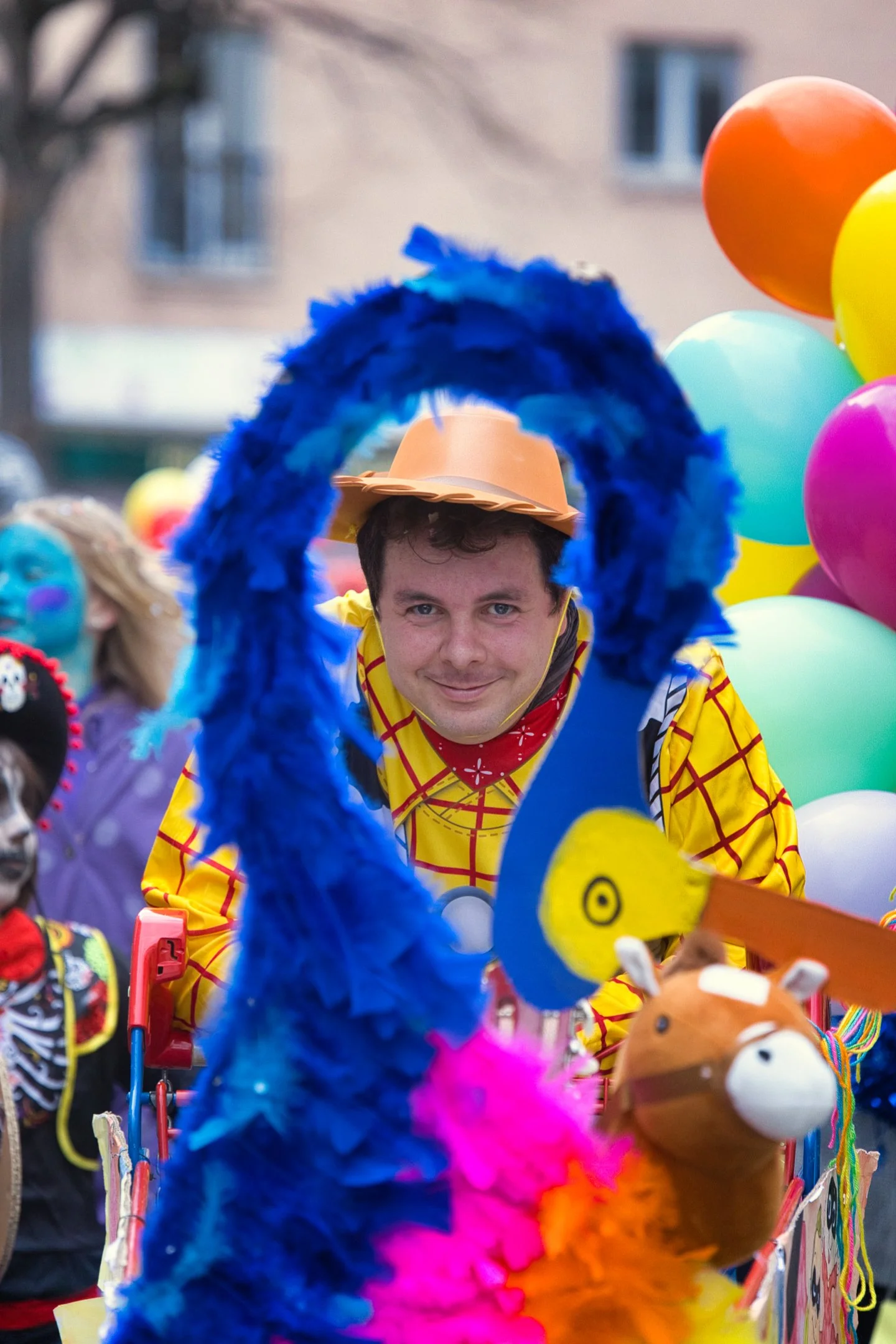 Un homme souriant déguisé en Woody de Toy Story, vue à travers un décor coloré de fête avec des ballons, en costume de carnaval.