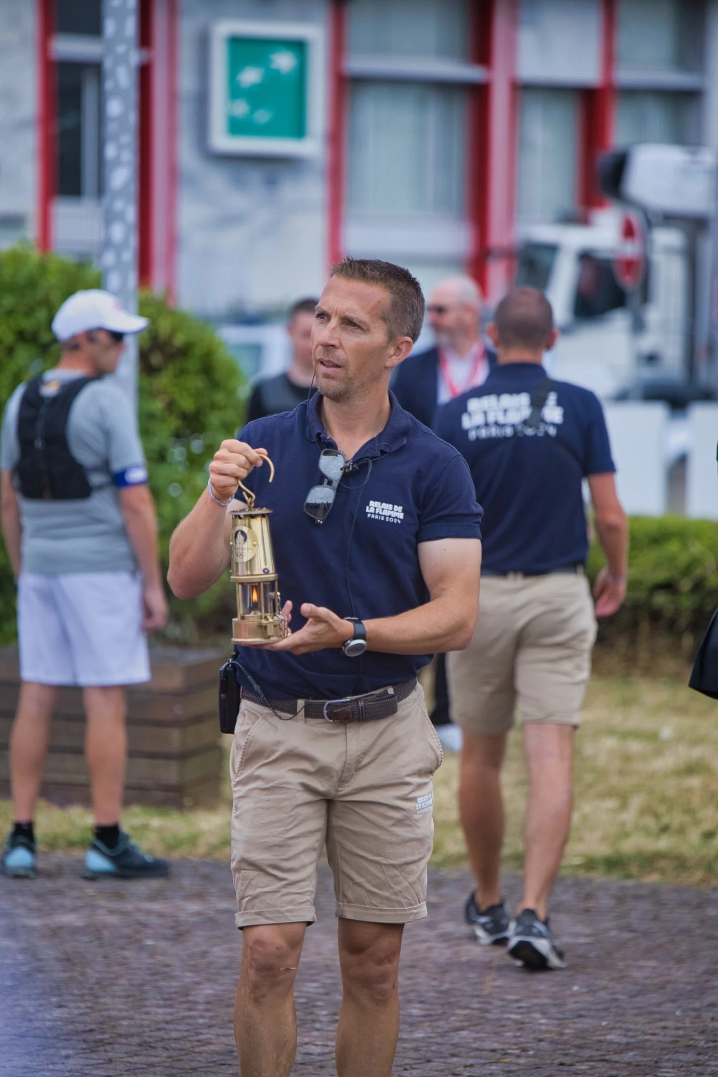 Un homme portant un polo bleu clair et des shorts beige, tenant une lanterne en métal lors d'un événement en extérieur. JO Paris 2024
