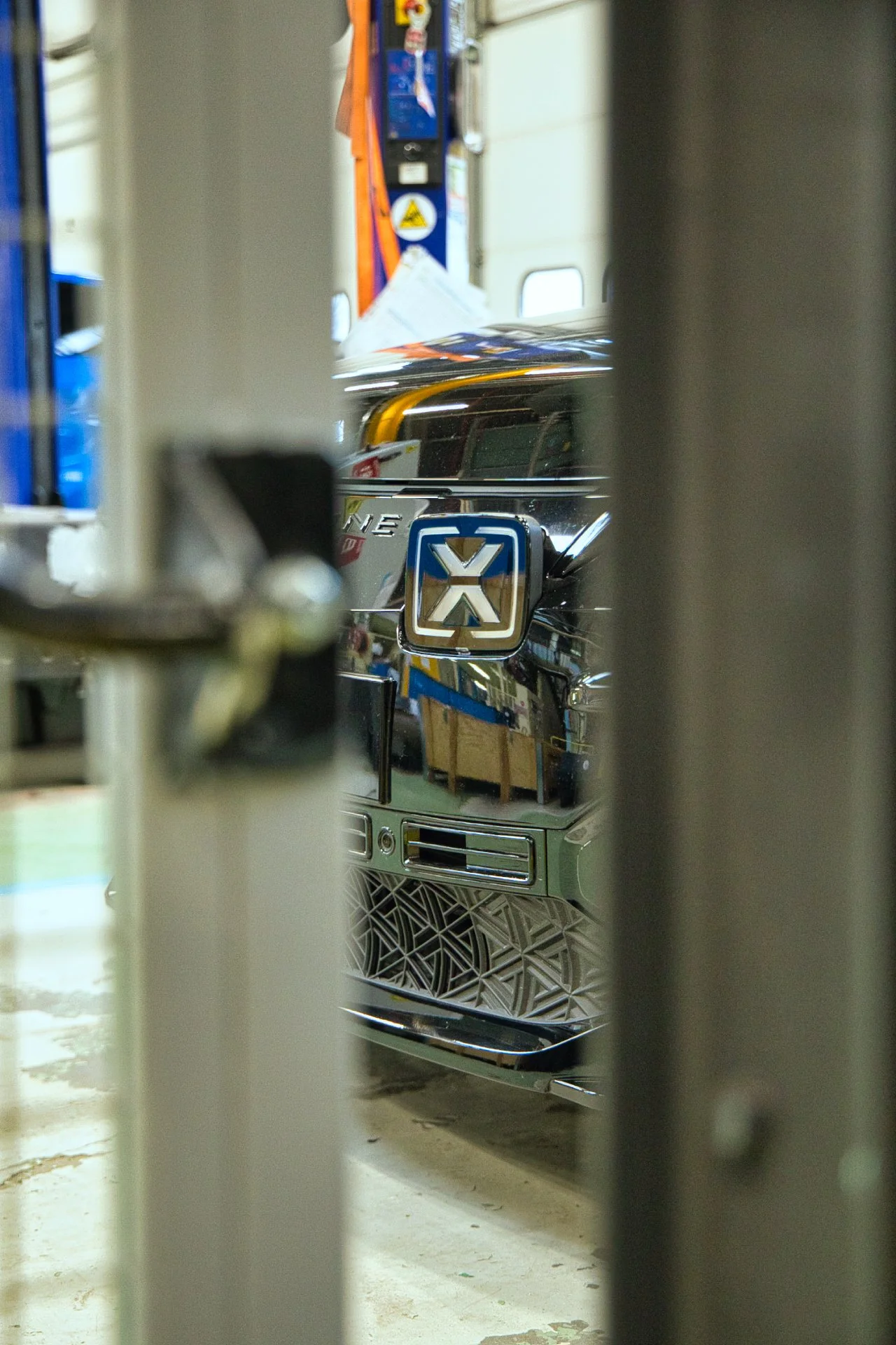 Une vue partielle de la face avant d'e l'Alpine A290, vue à travers une grille ou une porte, avec un badge ou logo en relief au centre. Ampere groupe Renault 