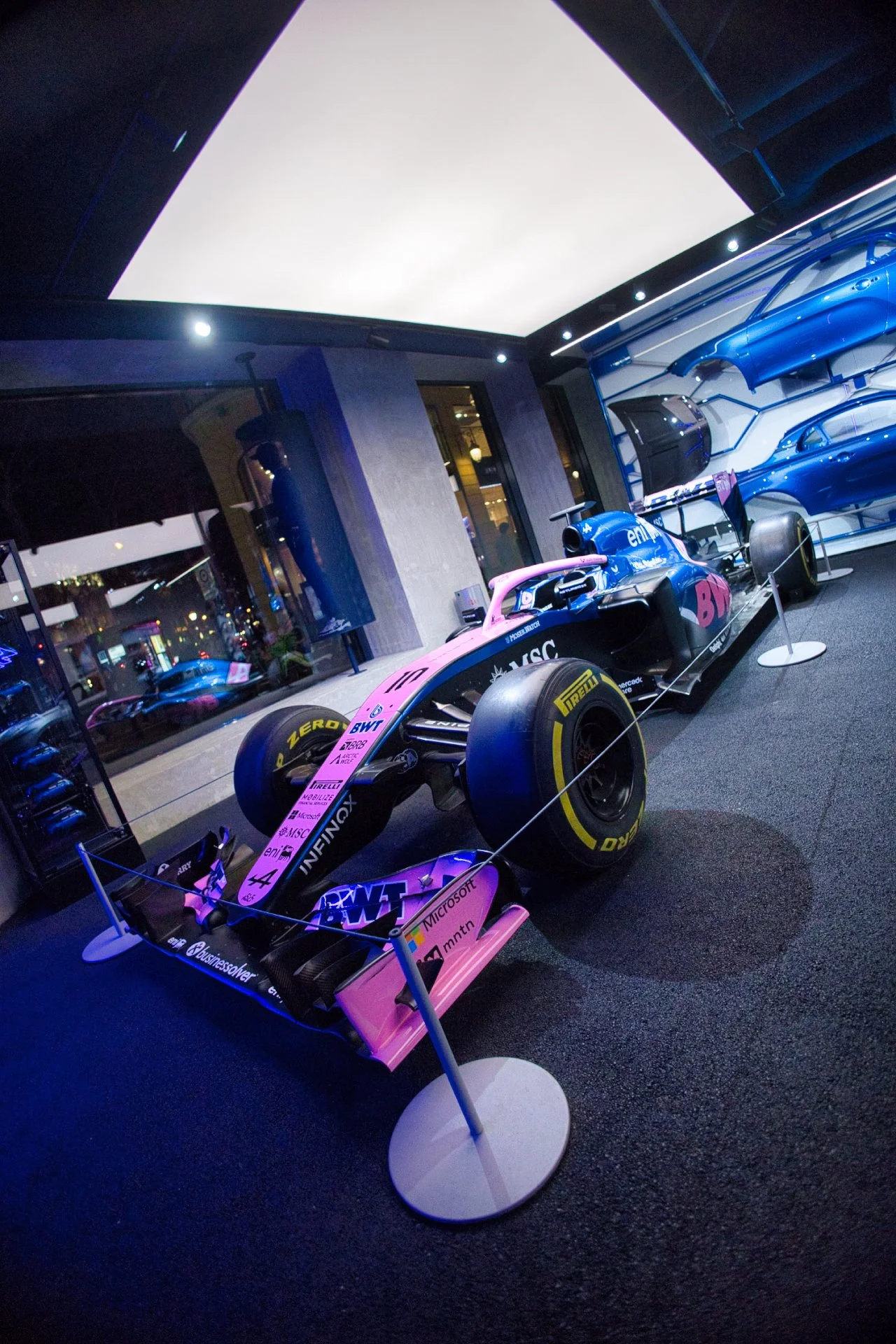Une voiture de Formule 1 rose et noire exposée dans une vitrine avec des pièces de voiture bleues en arrière-plan à l'atelier Alpine à Paris
