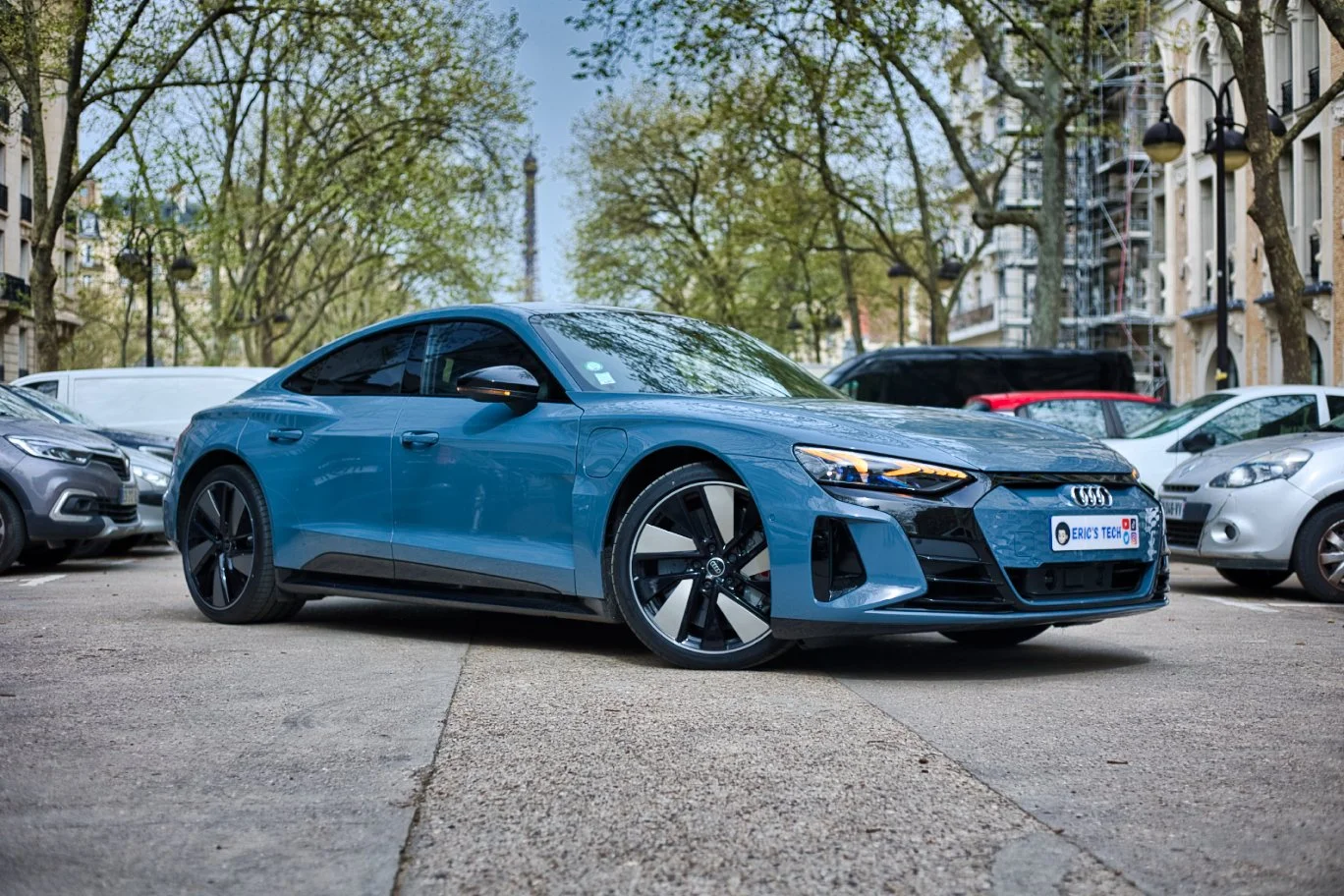 Audi e-tron rs bleue garée dans une rue urbaine, entourée d'autres véhicules, avec des arbres en arrière-plan et des bâtiments résidentiels.