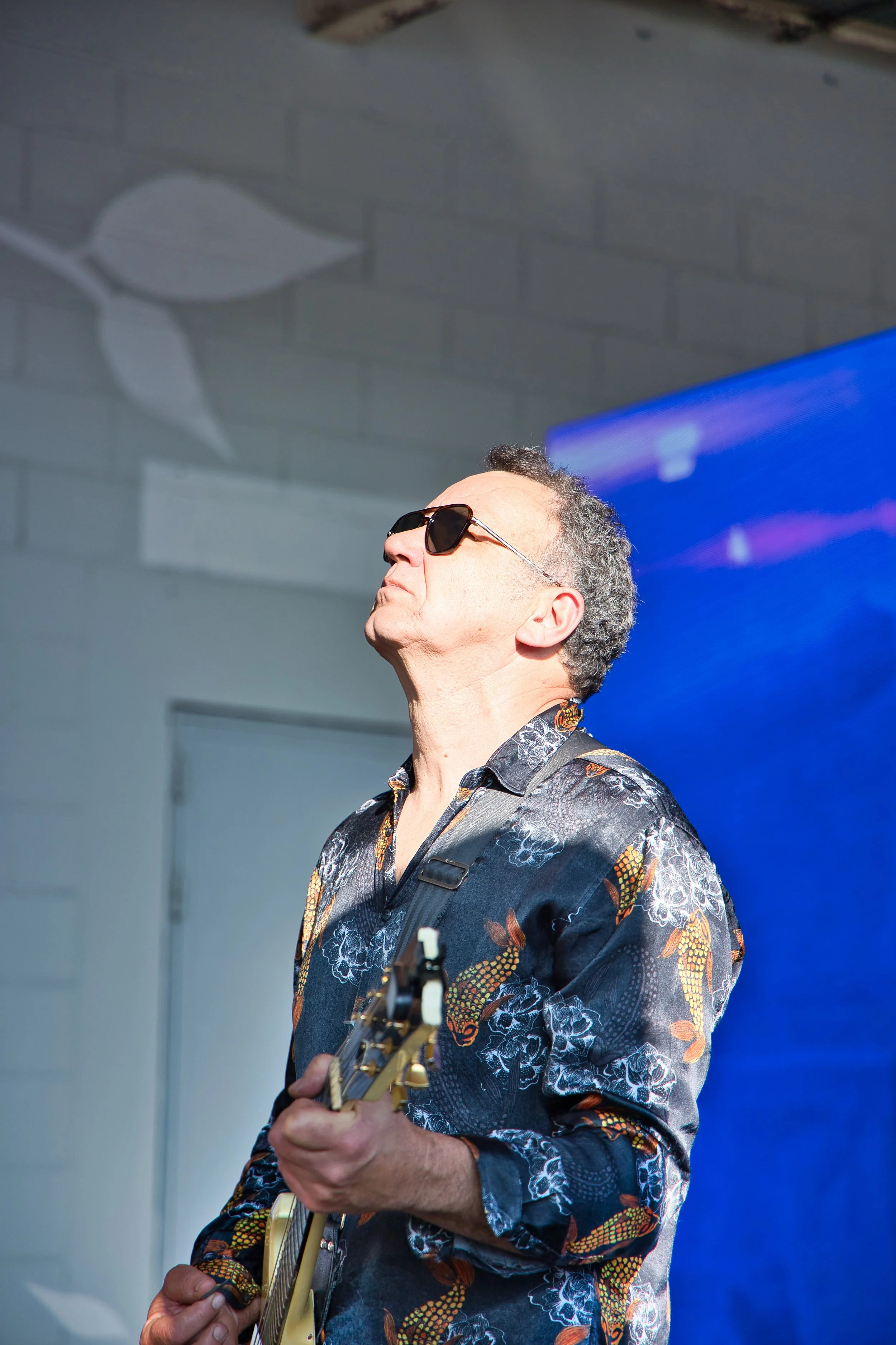 Un homme portant des lunettes de soleil et une chemise à motifs joue de la guitare devant un mur blanc avec un dessin de feuille, en intérieur.